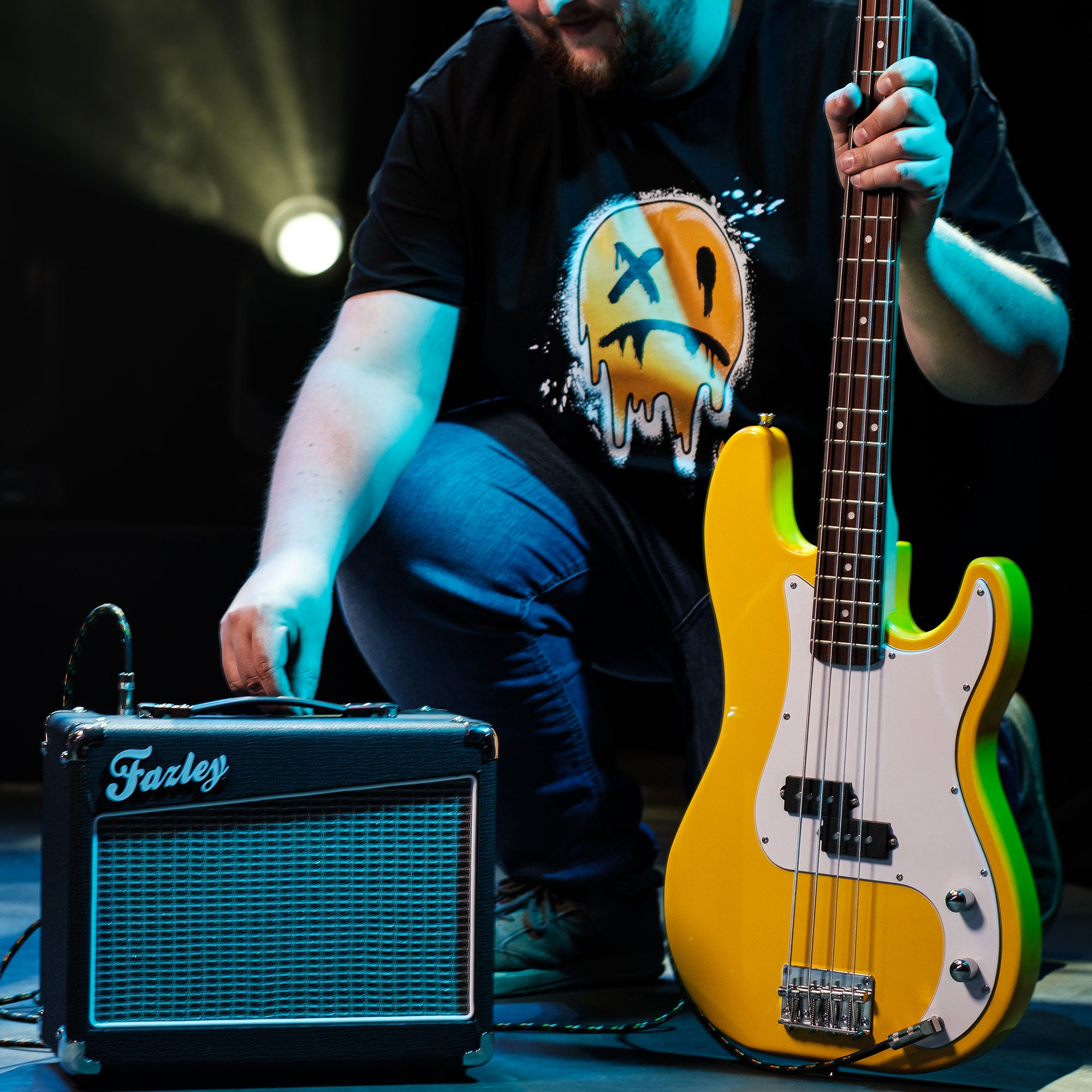 Een man knielt naast een gele basgitaar en een versterker.