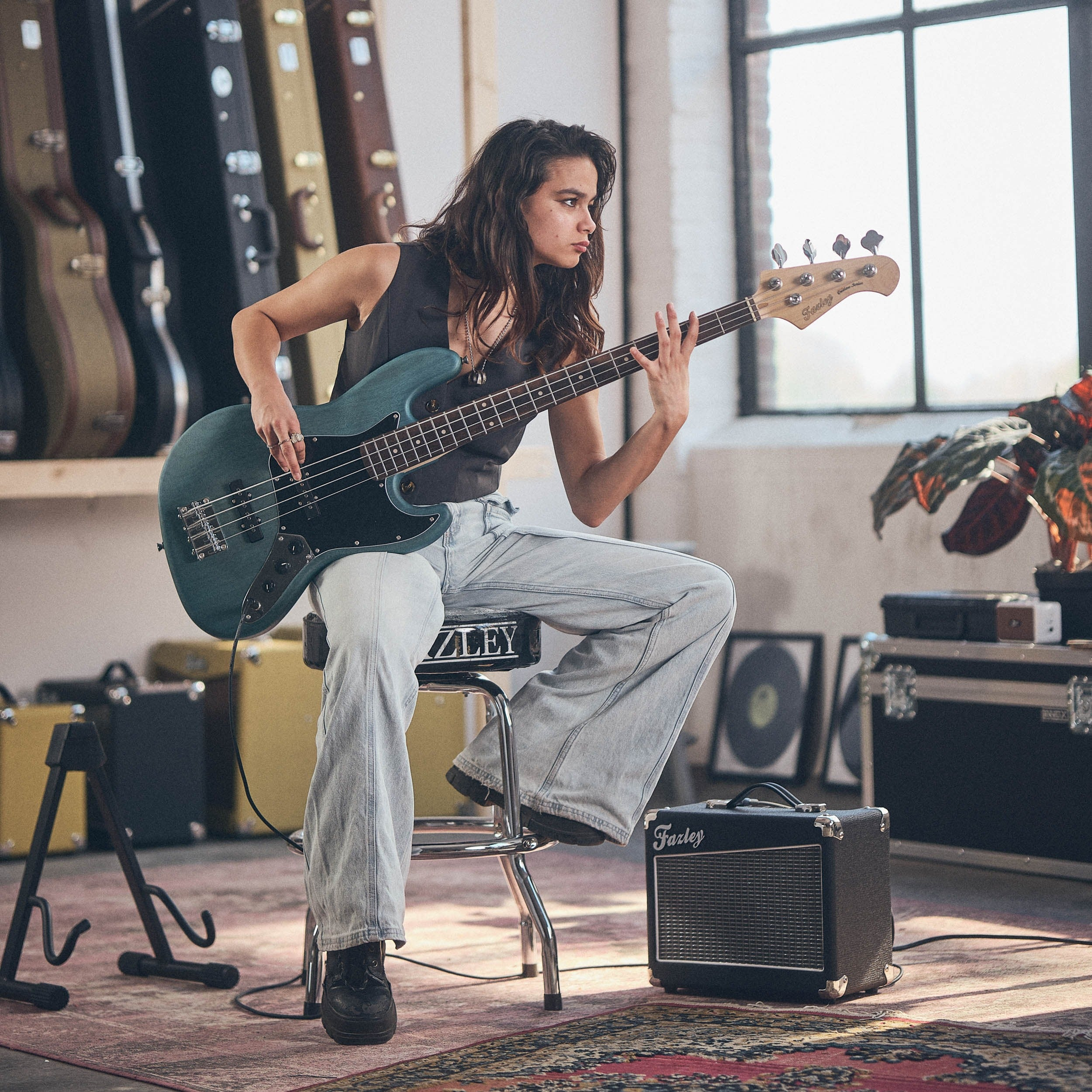Vrouw speelt op een teal basgitaar in een kamer met gitaarkoffers en versterkers.