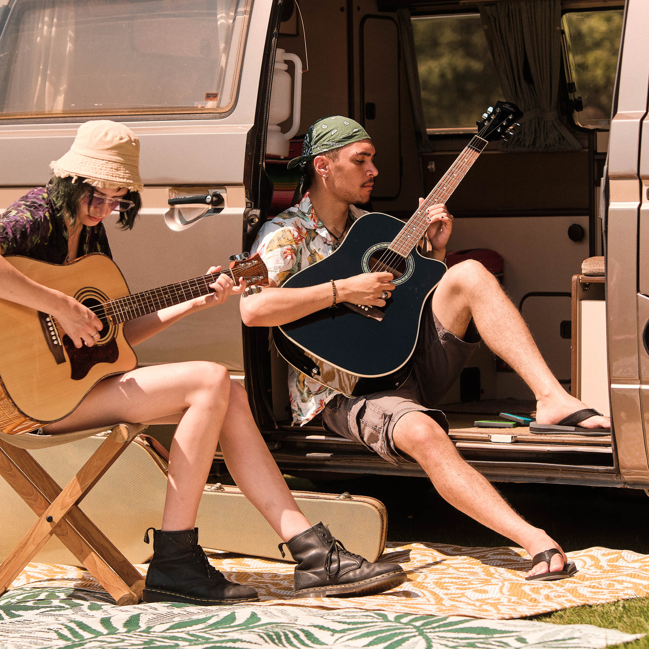 Two people play guitars by a van on a blanket. One person sits on a chair.