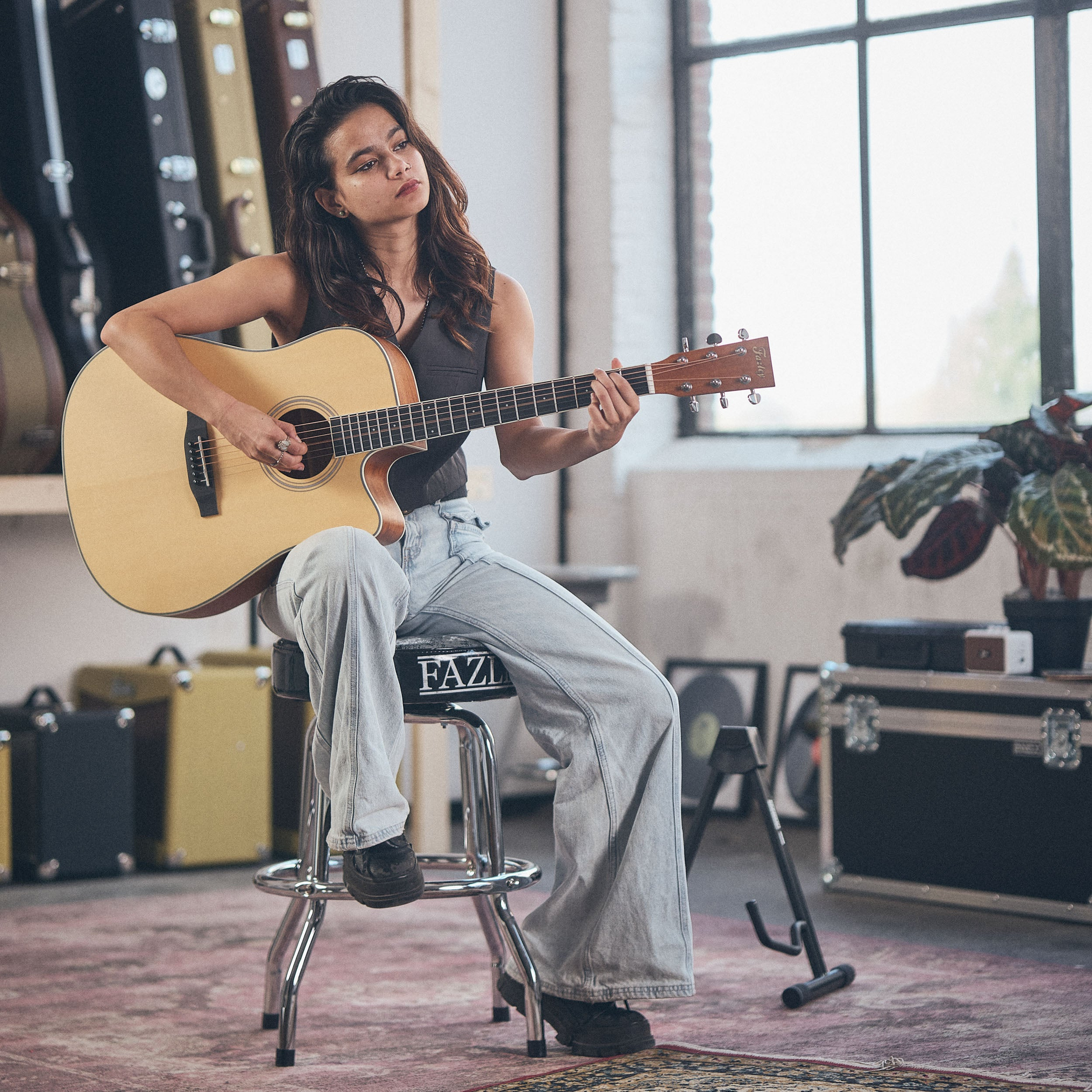 Een vrouw speelt akoestische gitaar, zittend op een kruk. Gitaren op de achtergrond.