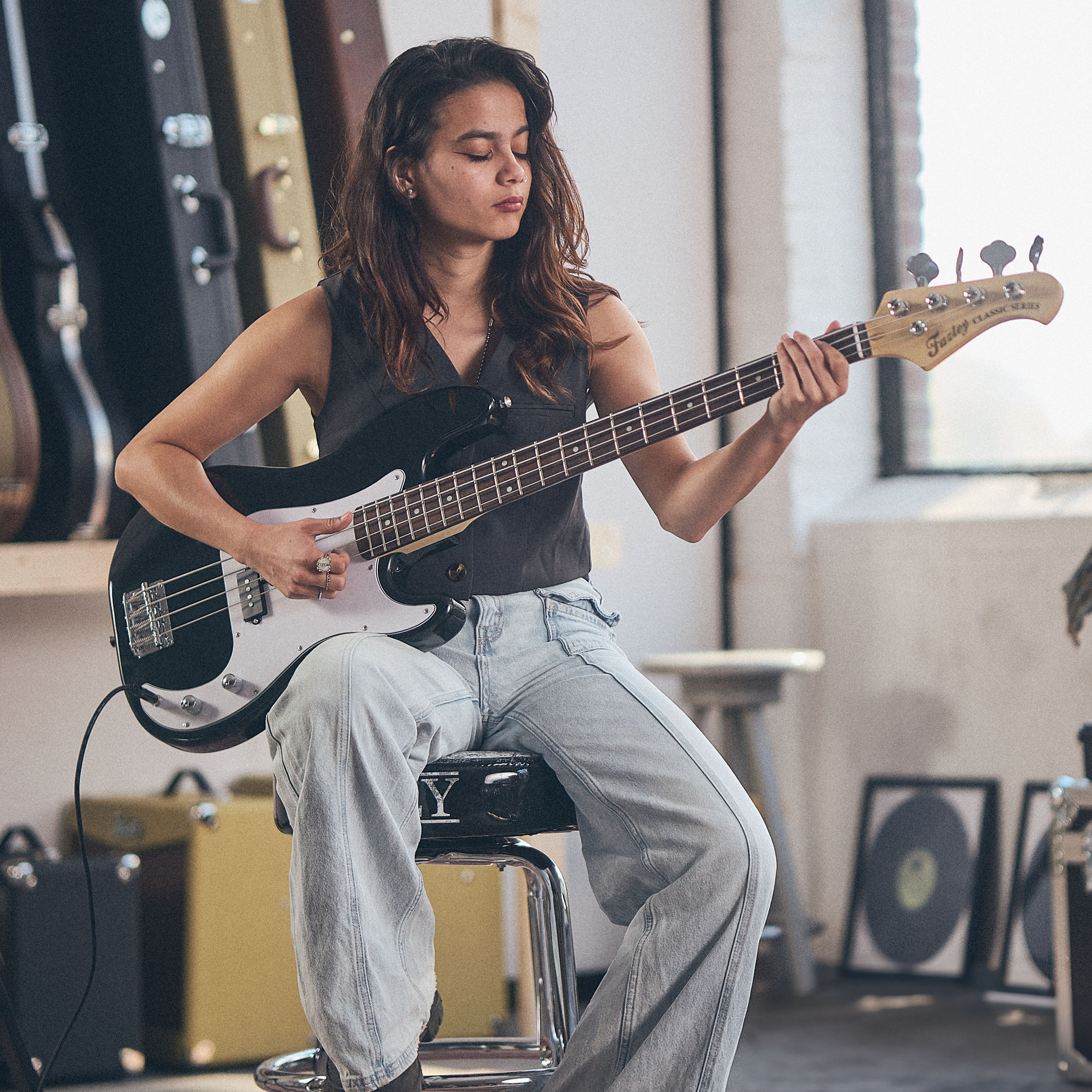 Een vrouw speelt op een zwarte elektrische basgitaar zittend op een kruk.