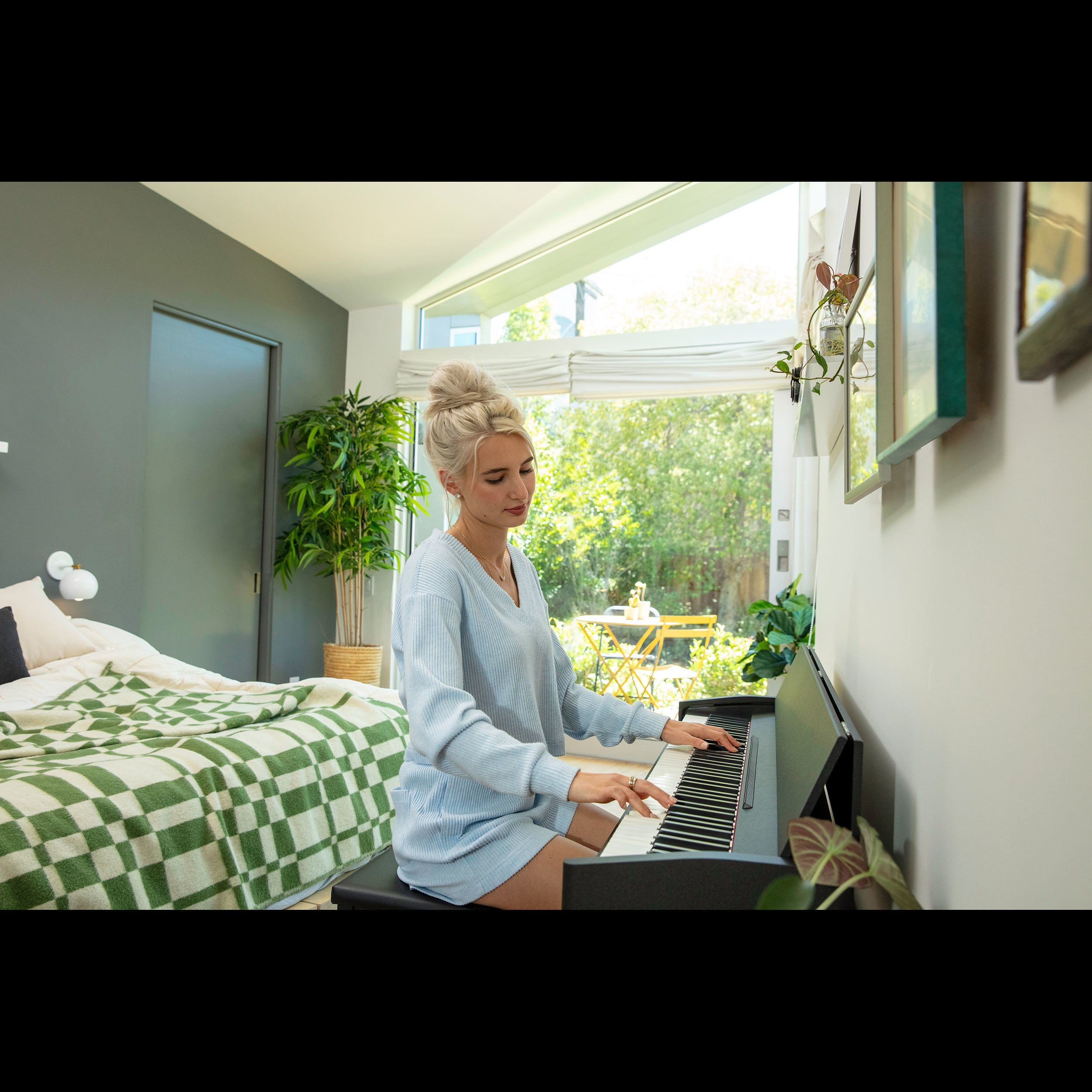 A woman plays the piano in a bright room overlooking greenery.