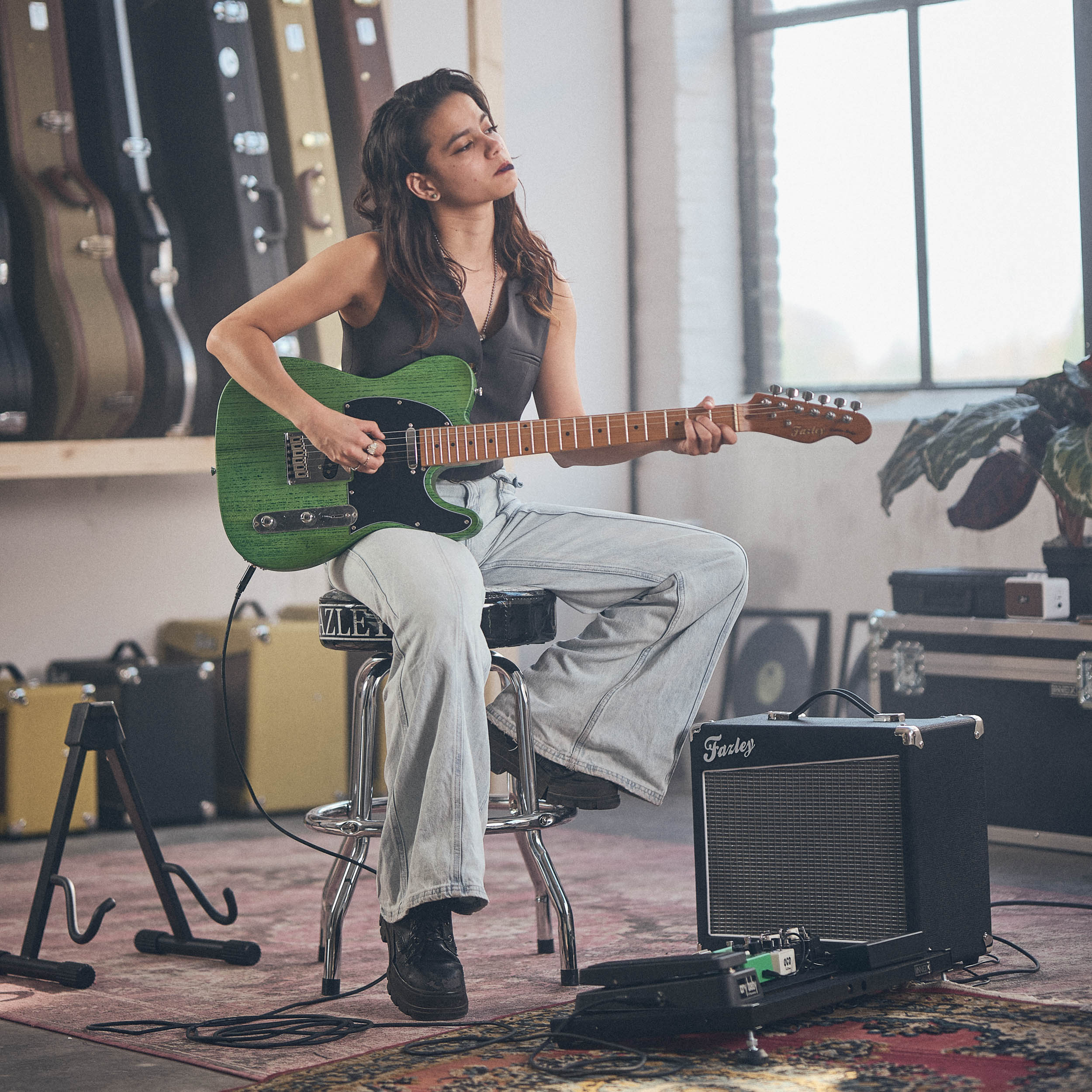 Vrouw speelt groene elektrische gitaar, zit op kruk, versterker en gitaarkoffers op achtergrond.