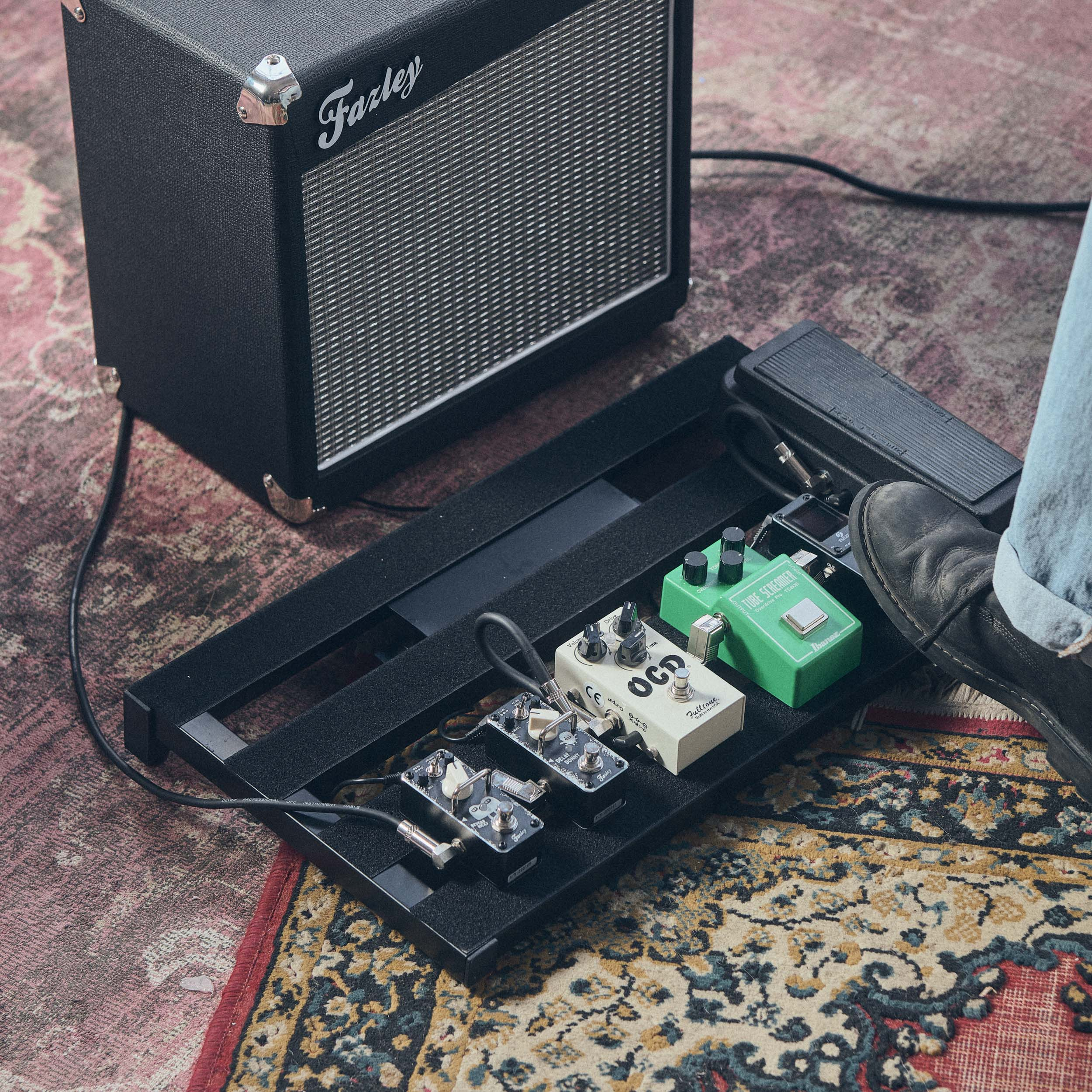 Een gitaarversterker met effectpedalen op een kleed. Een voet stapt op een pedaal.