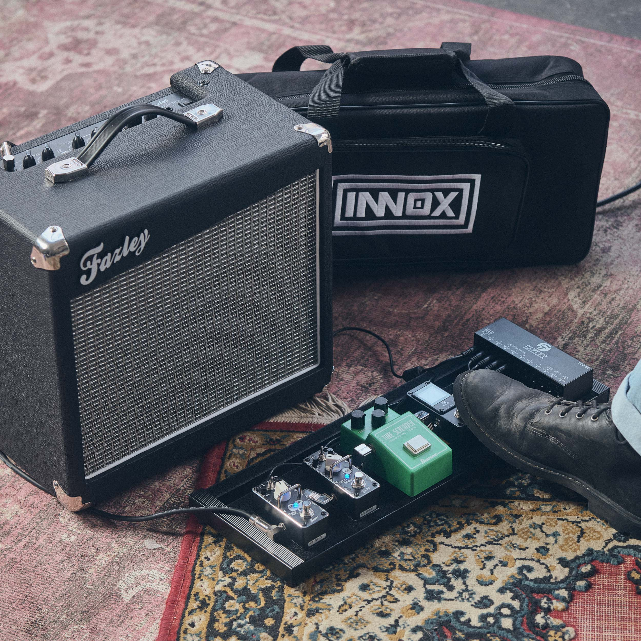 Een gitaarversterker, tas en pedalboard met effecten op een tapijt.