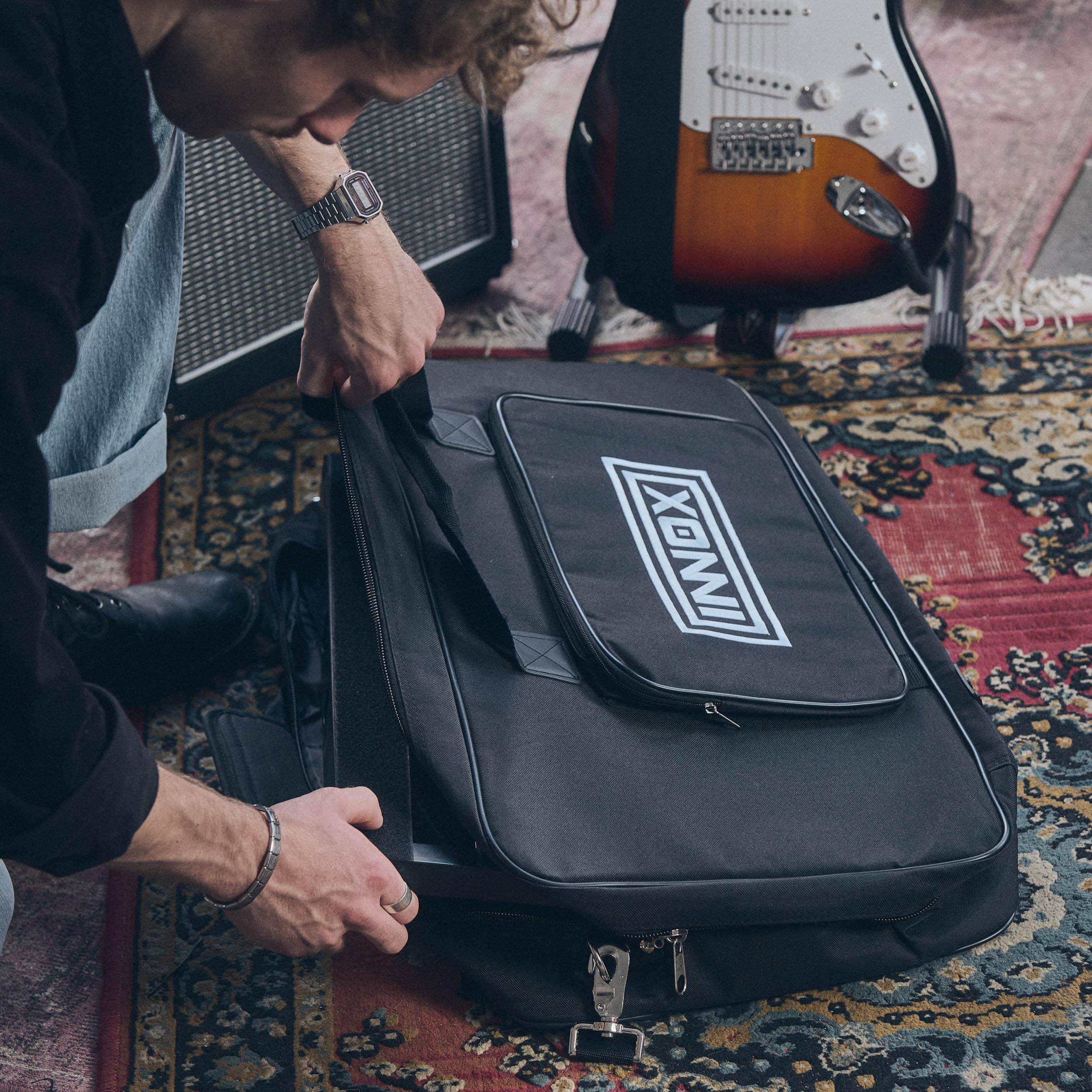 Man sluit zwarte tas met wit logo naast gitaar en versterker.