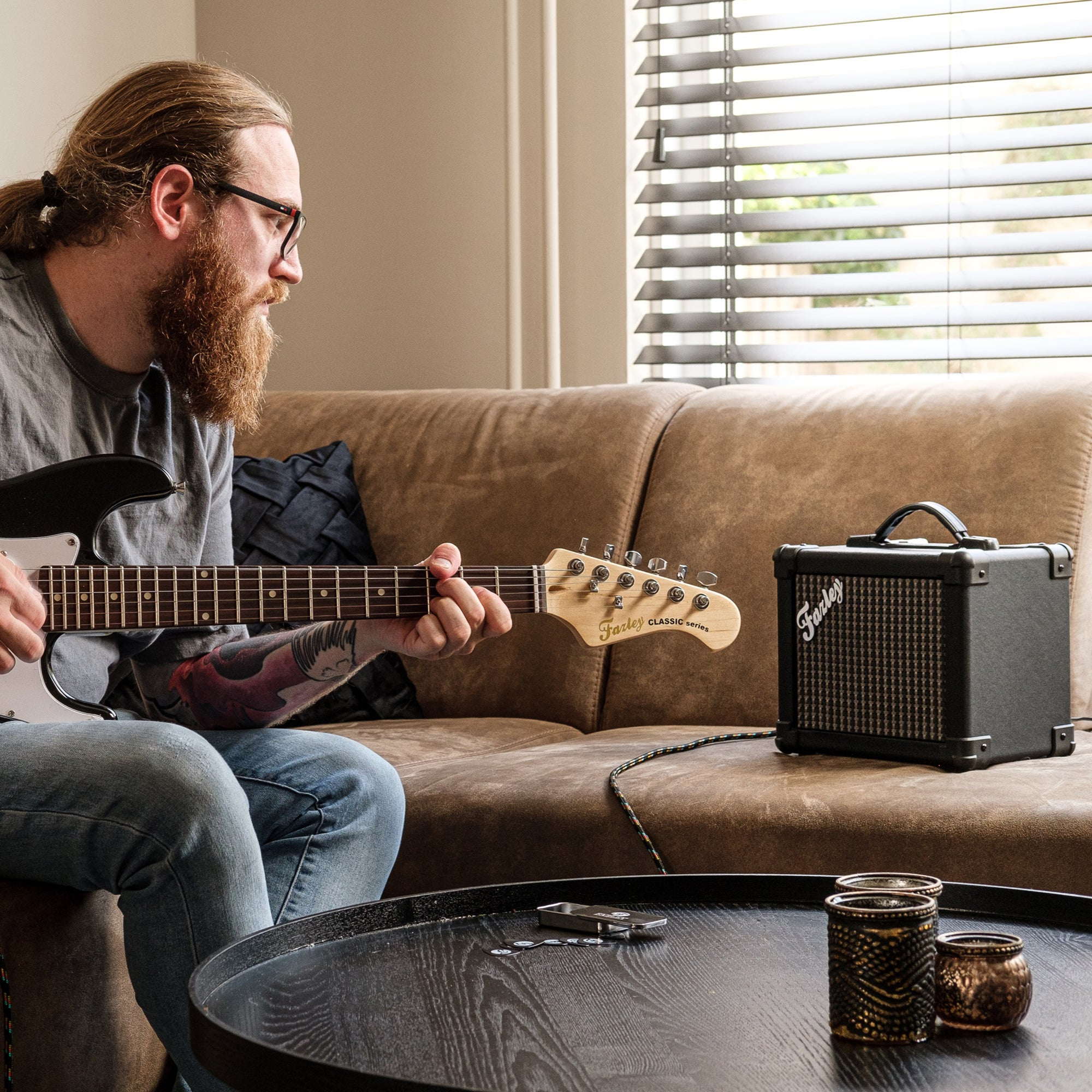 Man met baard speelt gitaar, versterker op bank, jaloezieën achtergrond.