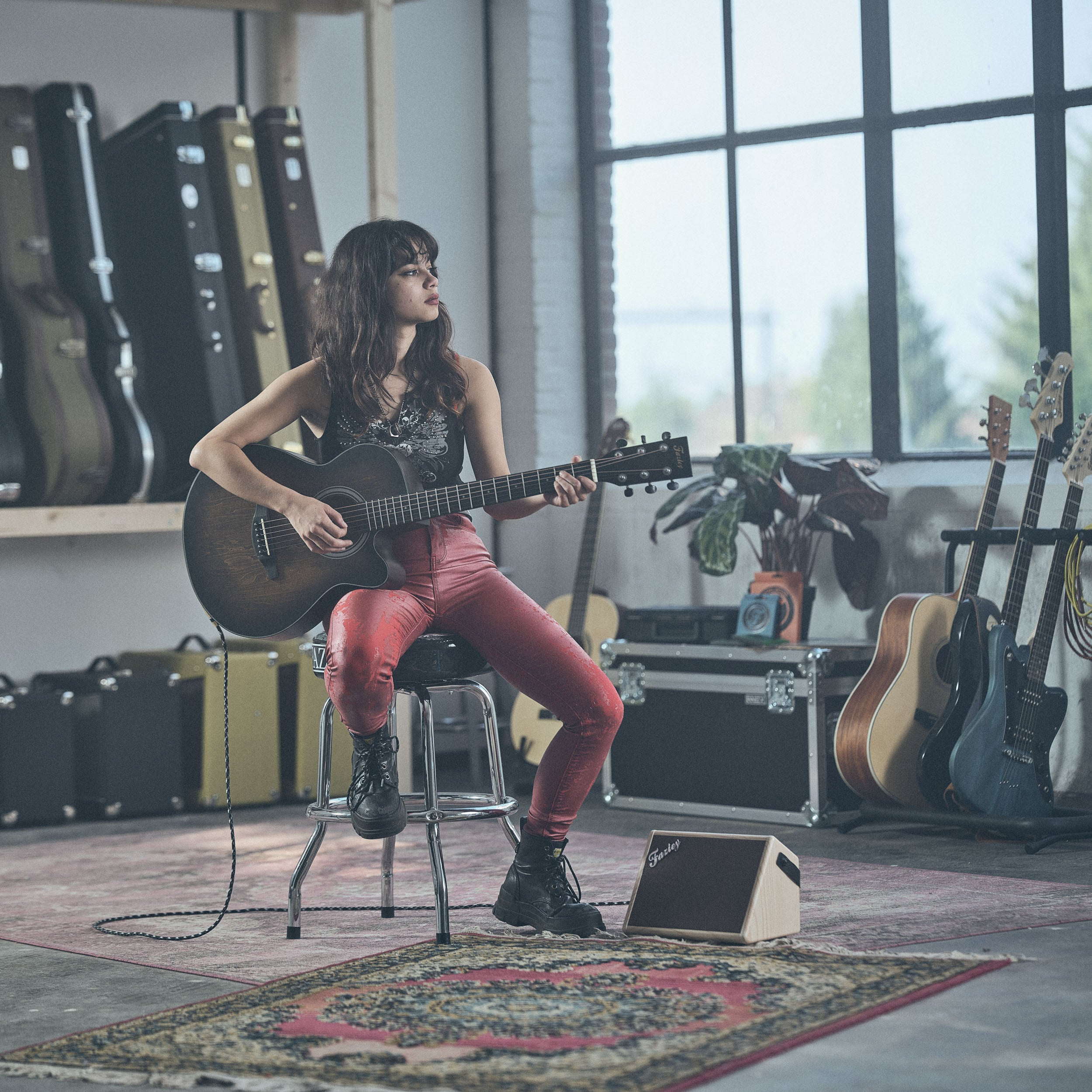 Een vrouw in een rode broek speelt akoestische gitaar in een kamer vol gitaren.