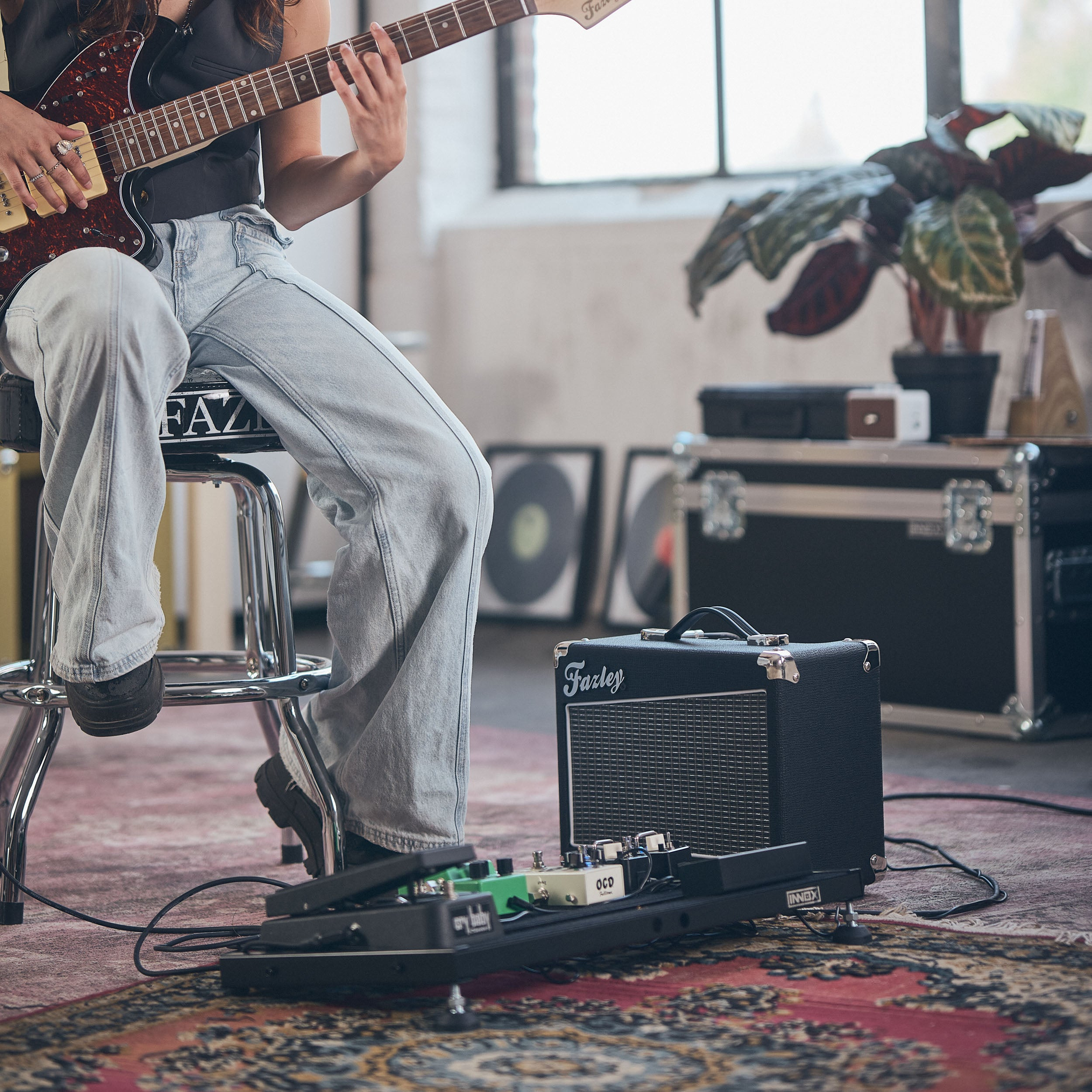 Een persoon speelt elektrische gitaar zittend op een kruk, naast versterker en pedalboard.