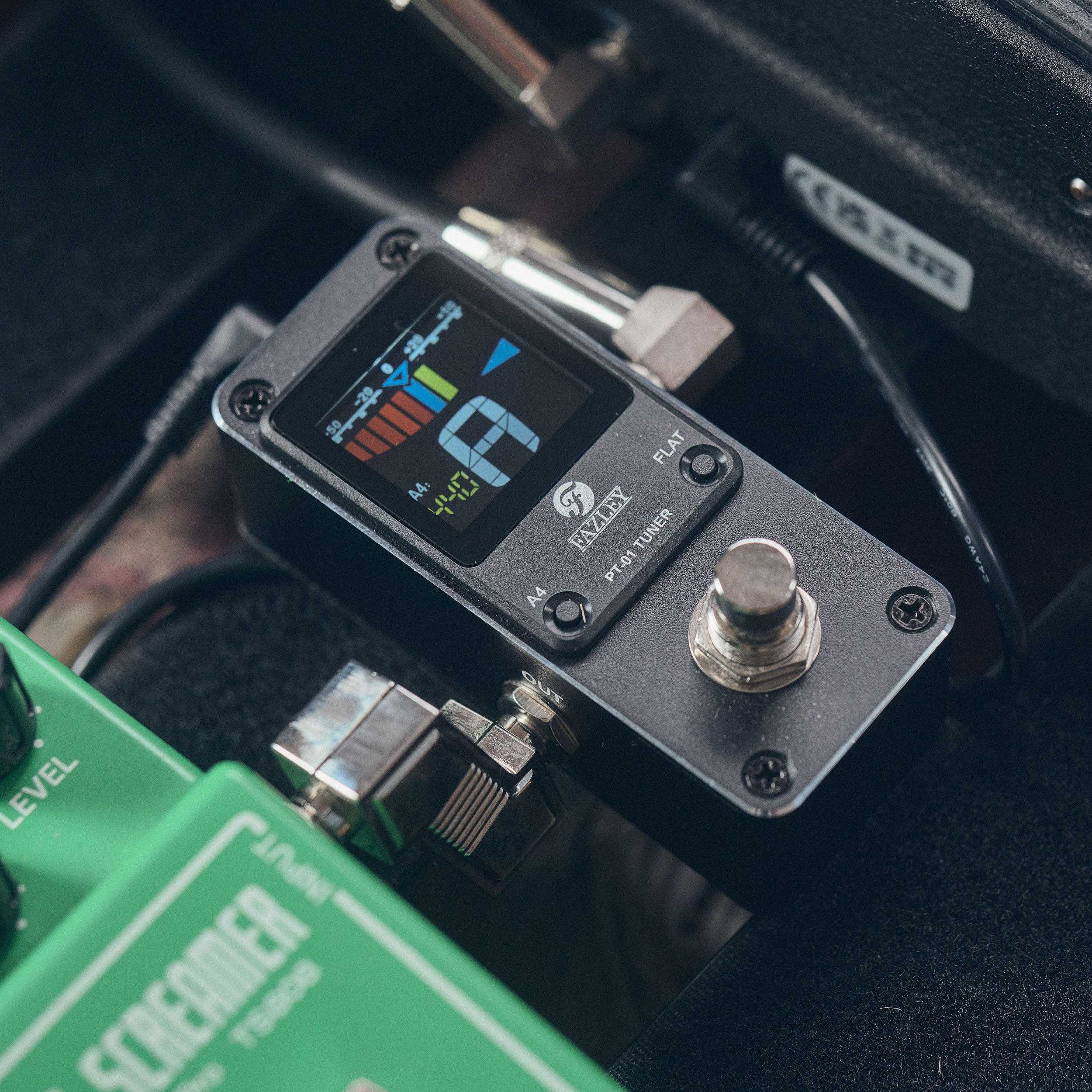 A black Fazley PT-01 tuner sits on a pedalboard next to a green effect pedal.