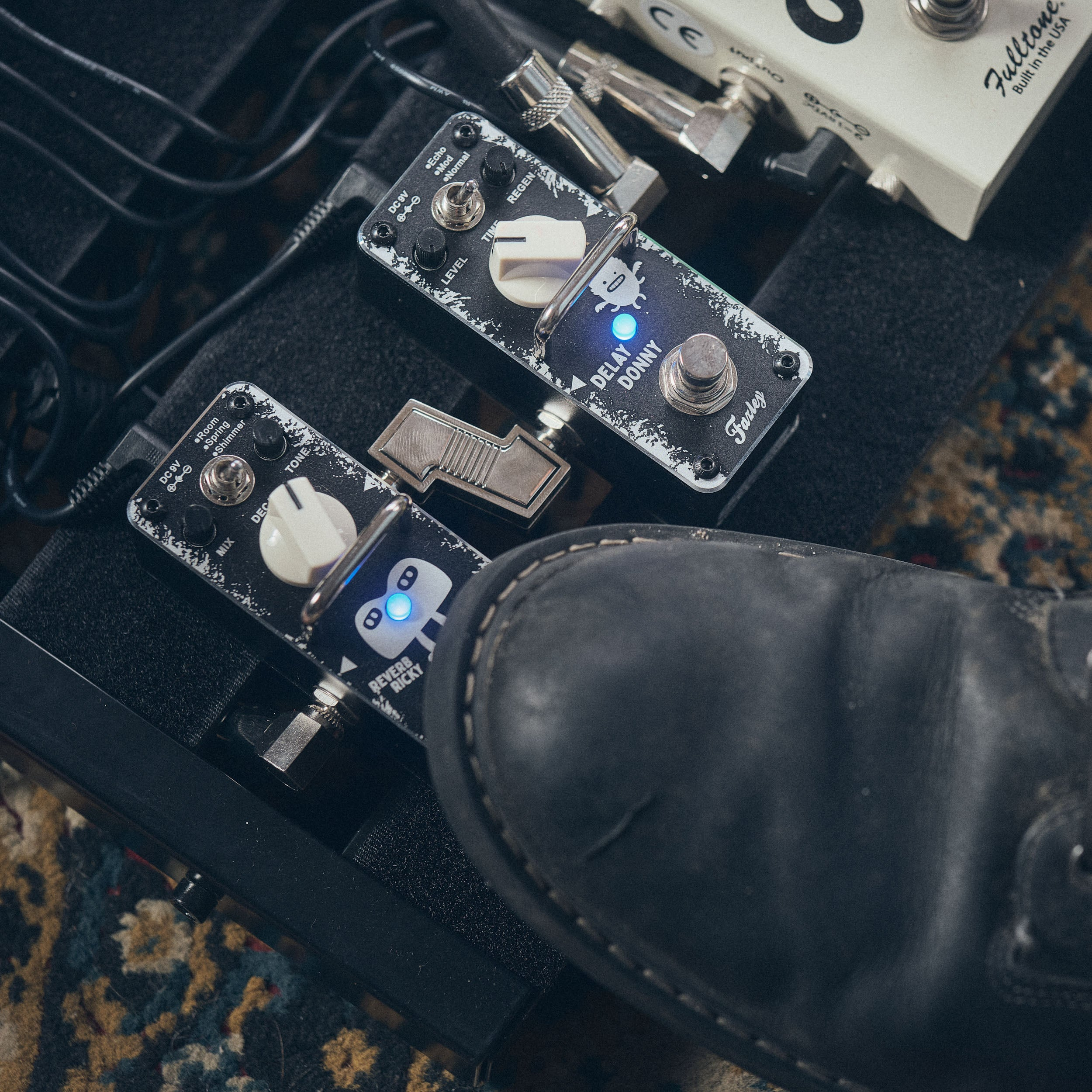 A black shoe steps on effect pedals on a pedalboard.