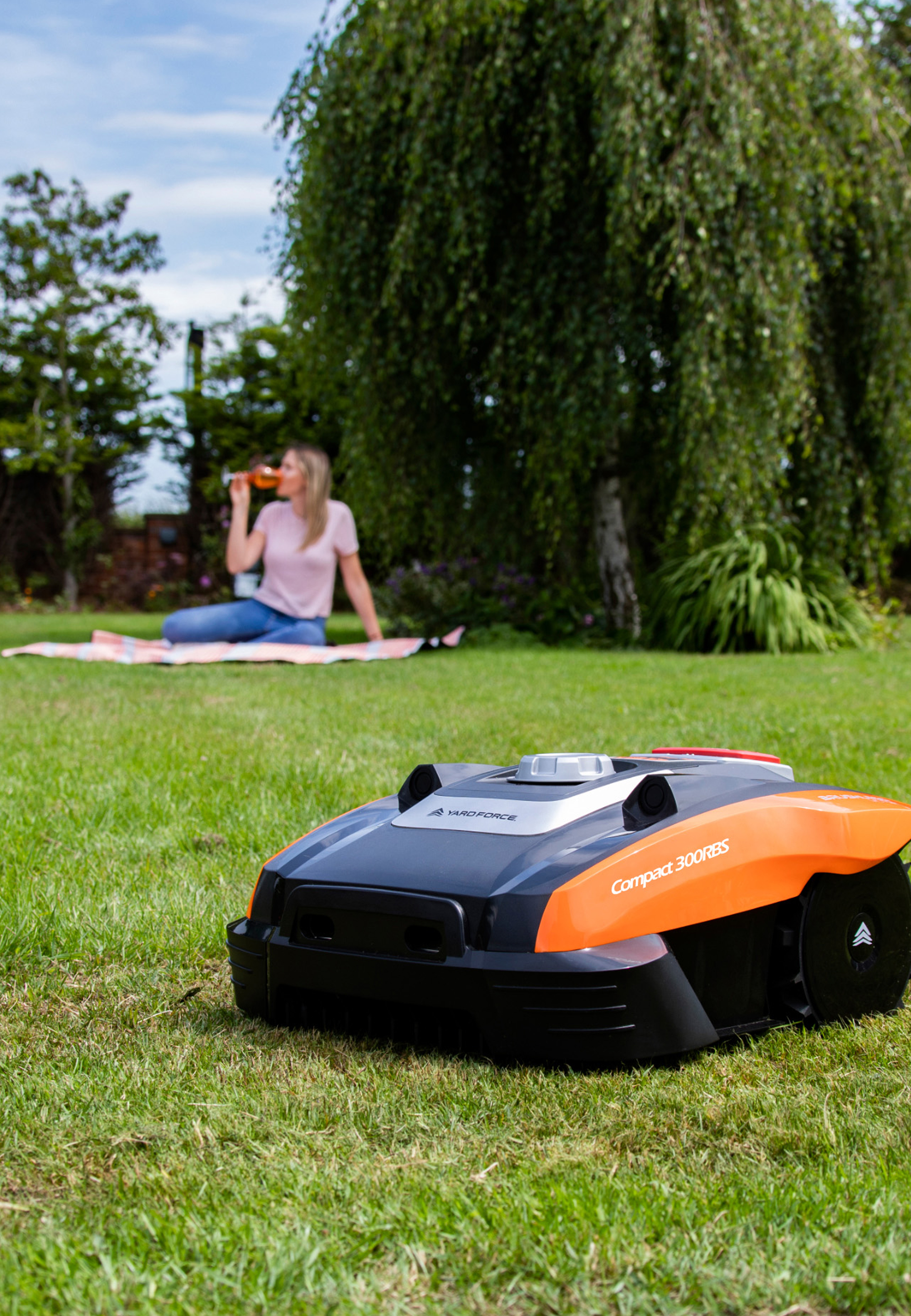 Ein Mähroboter steht auf einem Grasfeld, eine Frau picknickt im Hintergrund.