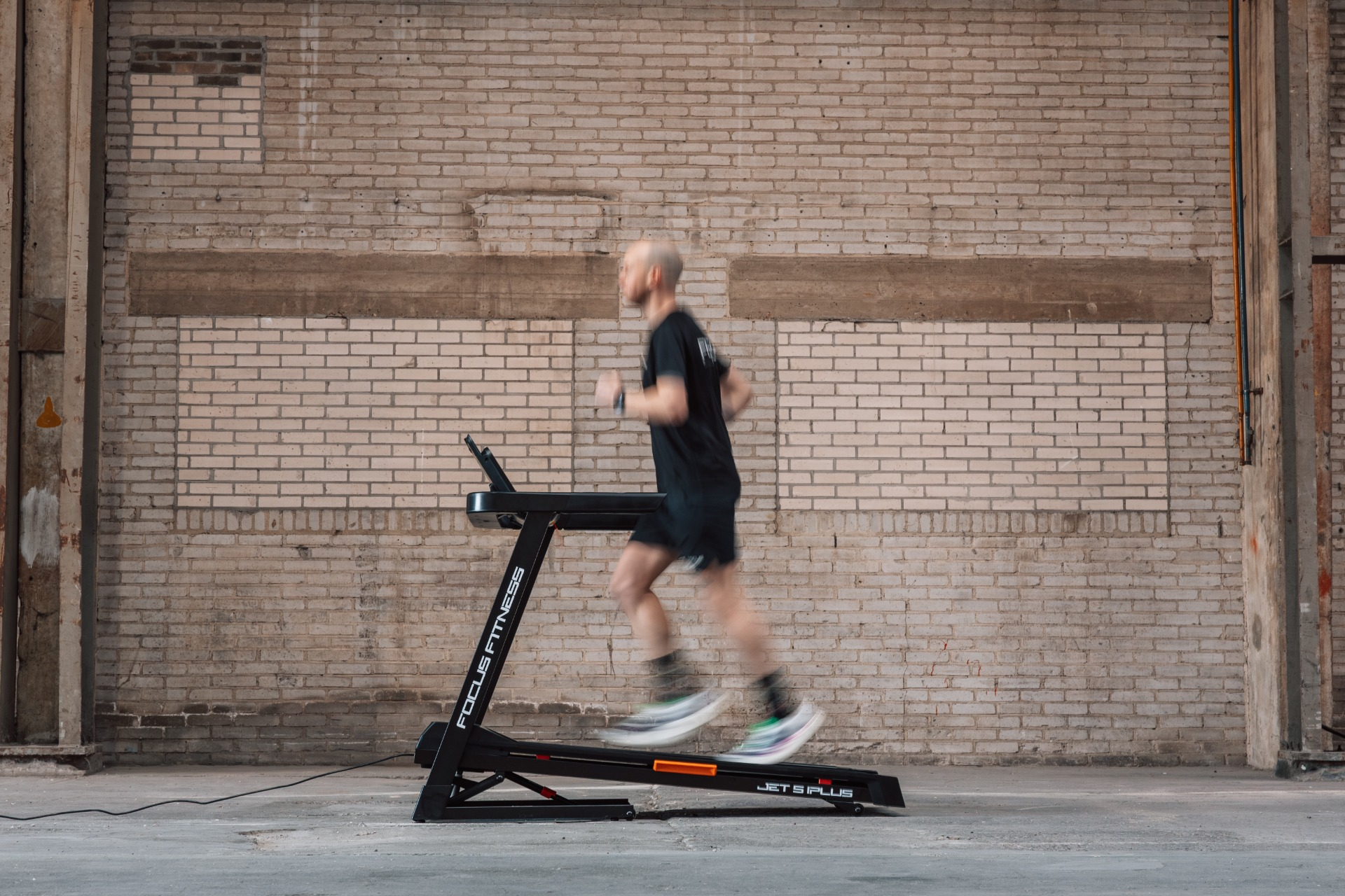 A man runs on a treadmill in front of a brick wall.