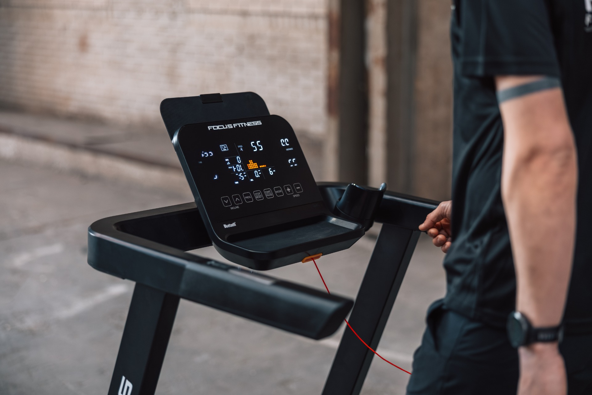 A man in black stands by a black treadmill. The display shows fitness data.