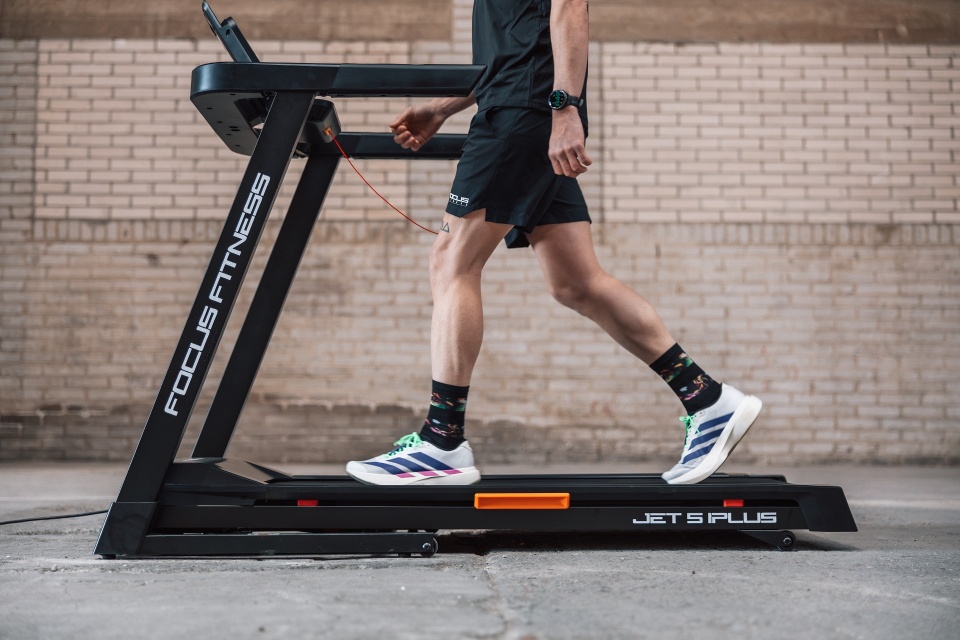 Person running on a black treadmill with a white brick wall in the background.