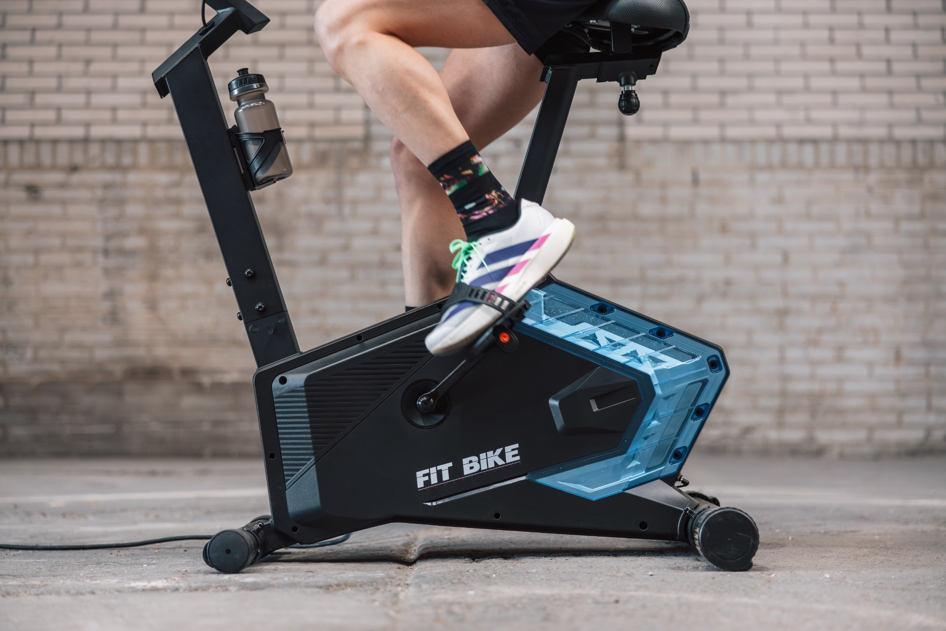 Persoon op een zwarte hometrainer met blauw detail, voor een bakstenen muur.