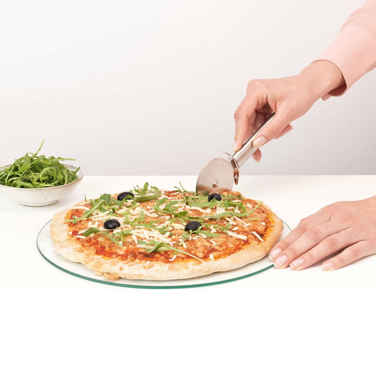 Eine Hand schneidet eine Pizza mit Rucola und Oliven mit einem Pizzaschneider.
