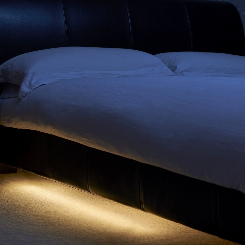 Una cama oscura con sábanas y almohadas blancas, iluminada desde abajo.