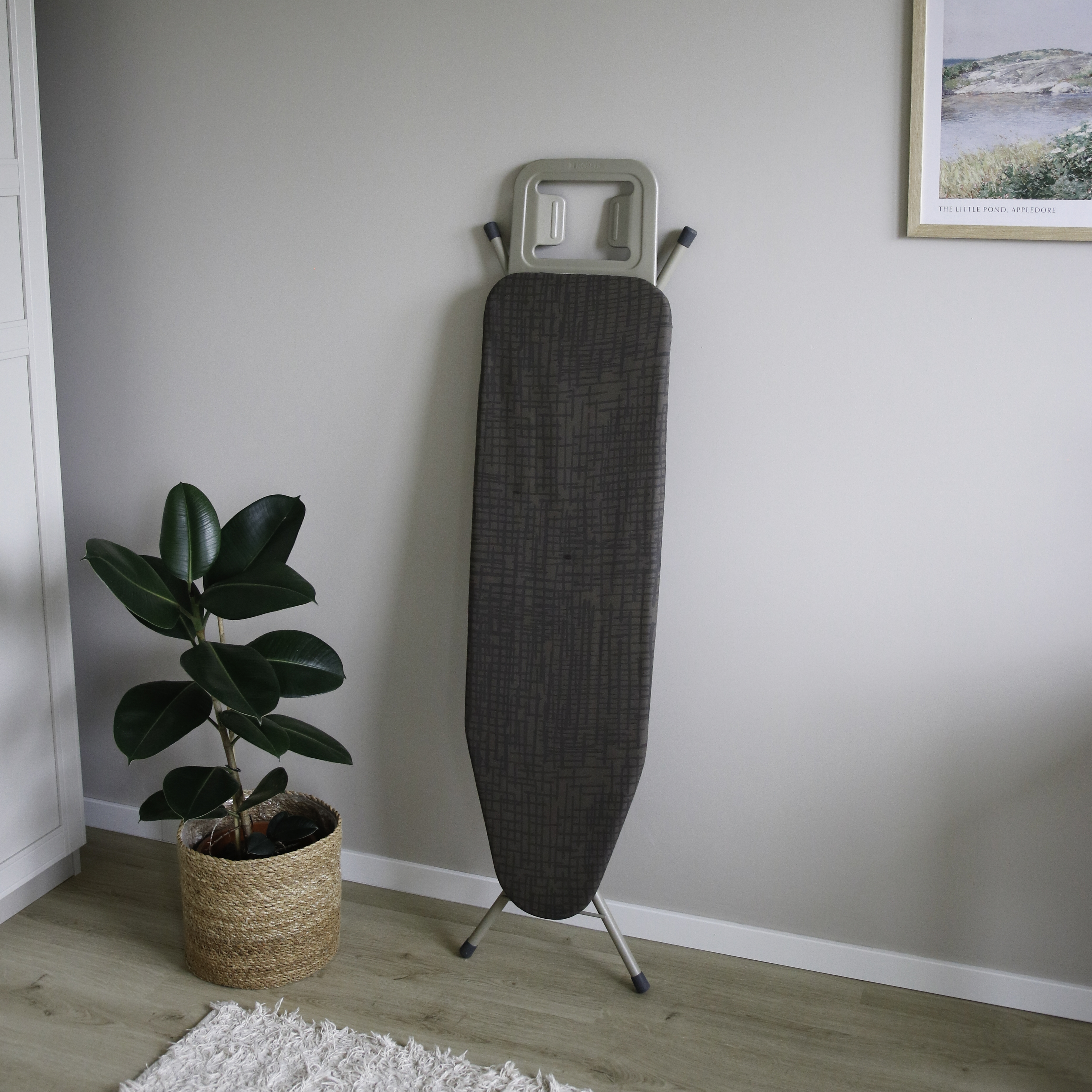 Une planche à repasser gris foncé est pliée contre un mur gris clair, à côté d'une plante.