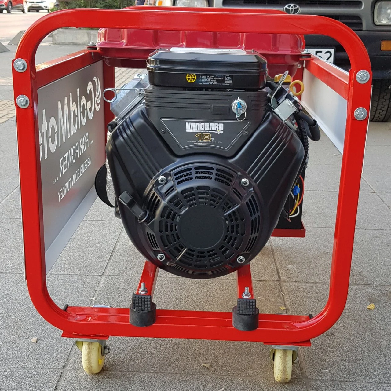 Siyah motorlu ve tekerlekli kırmızı bir jeneratör, kaldırımlı bir yolda duruyor.