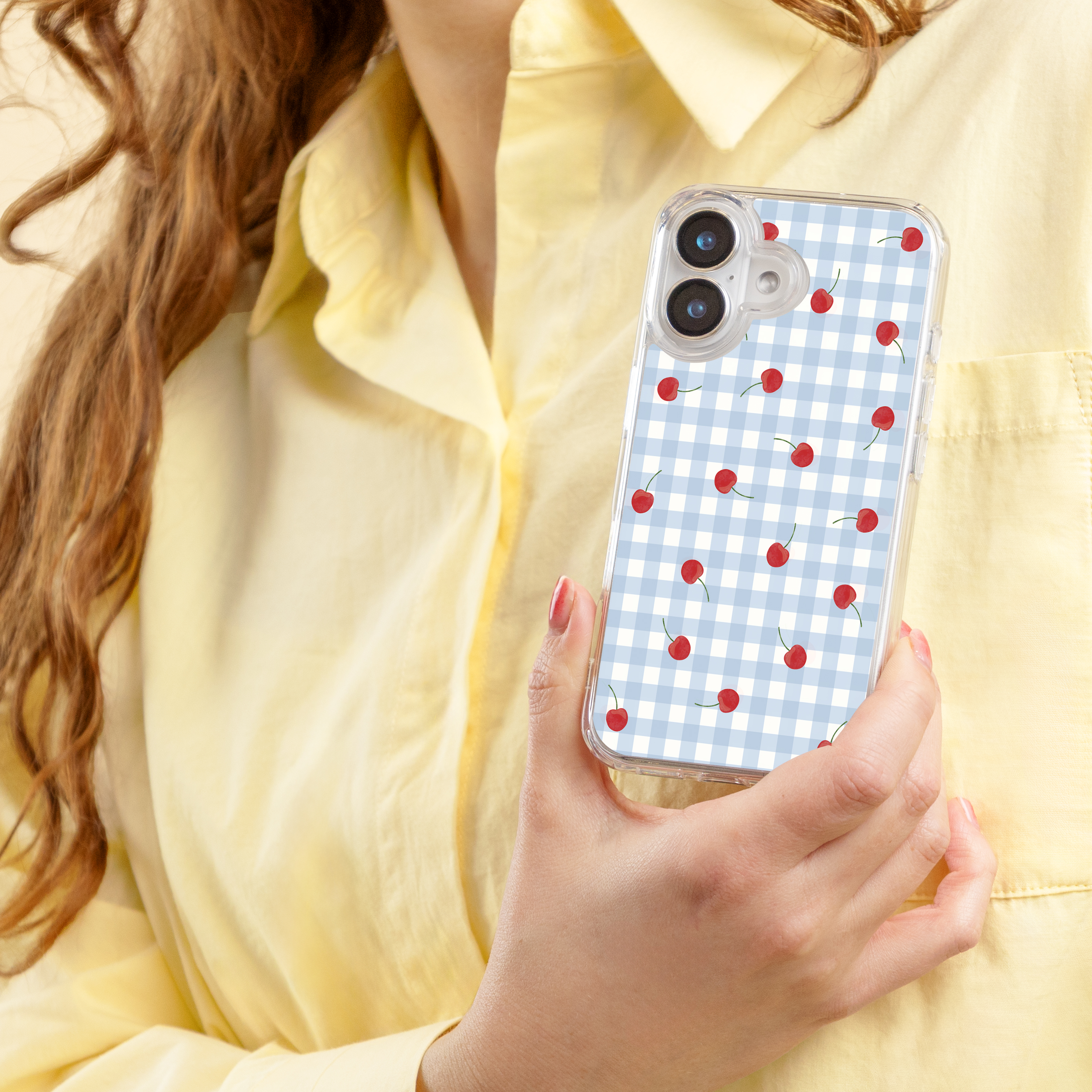 Een persoon in een geel shirt houdt een telefoon met kersen op een blauw geblokte achtergrond.