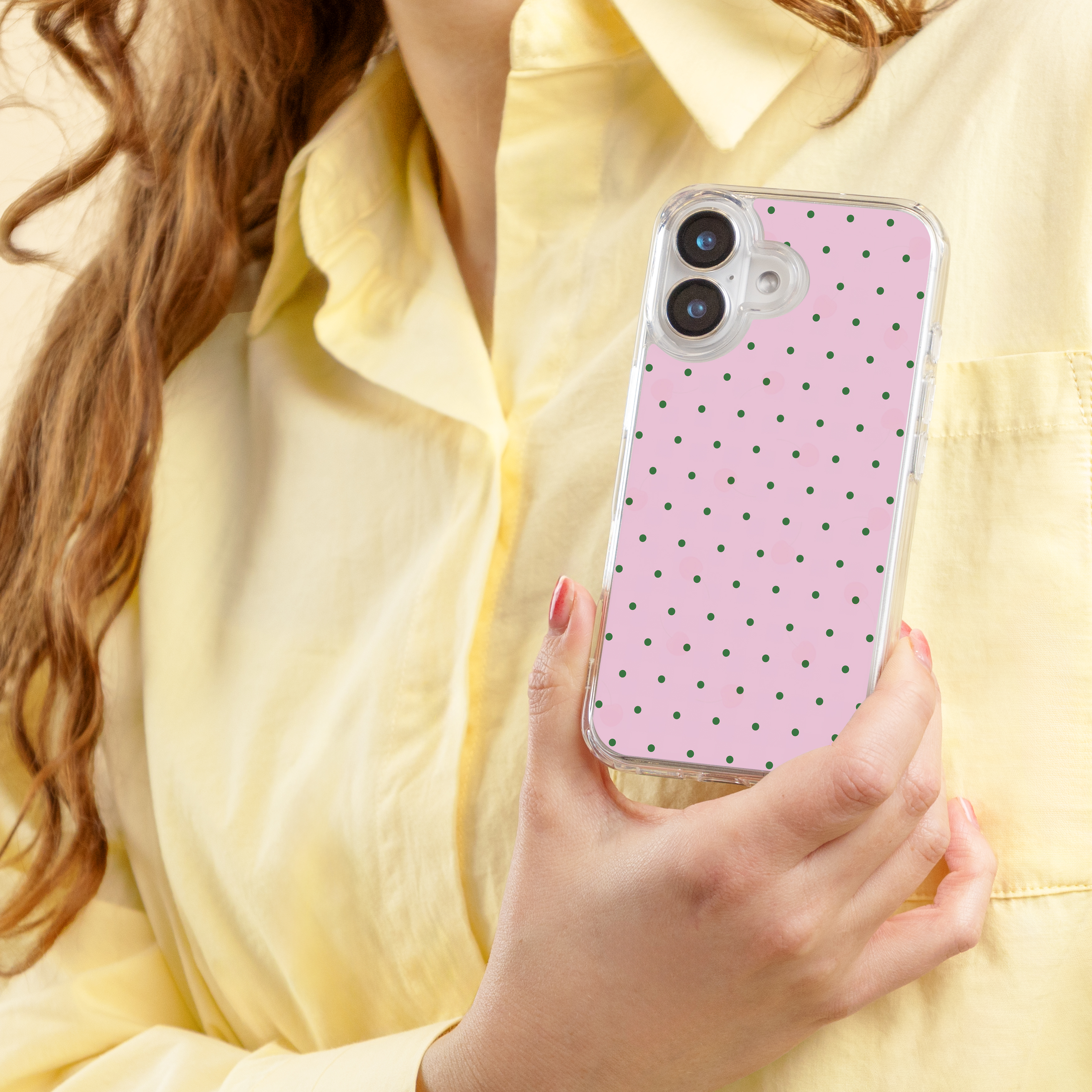Een persoon in een geel overhemd houdt een telefoon met roze polkadot hoesje vast.