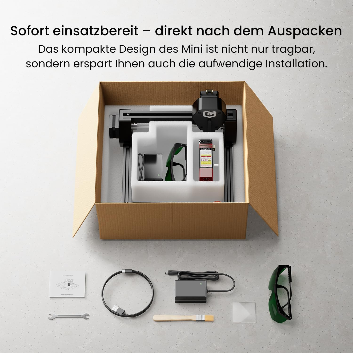 Ein Lasergravierer und Zubehör liegen in einer geöffneten Box auf grauem Untergrund.