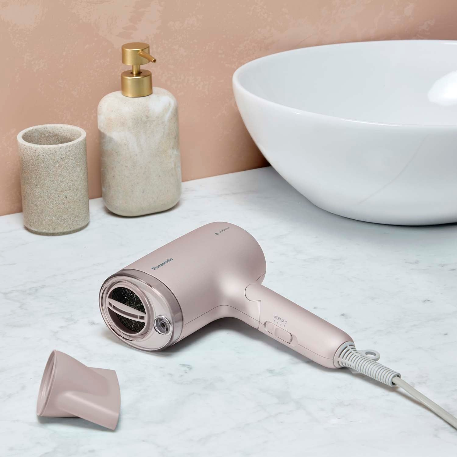 Un asciugacapelli color oro rosa poggia sul marmo accanto a un dispenser di sapone e un lavandino.