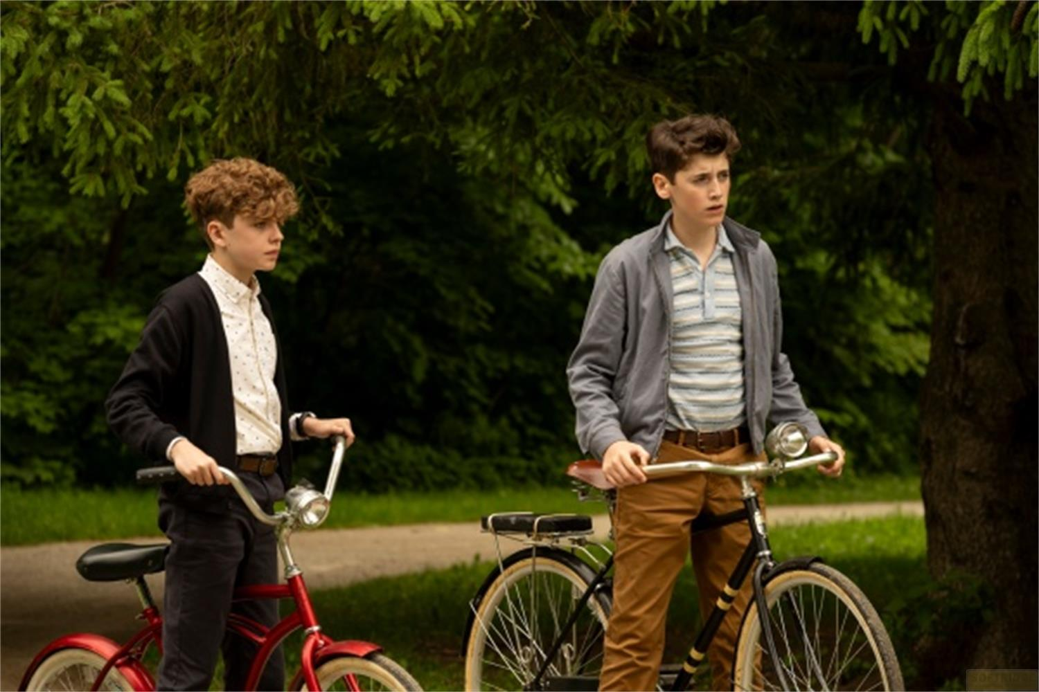 Due ragazzi stanno con le biciclette in un parco. Alberi verdi e fogliame li circondano.