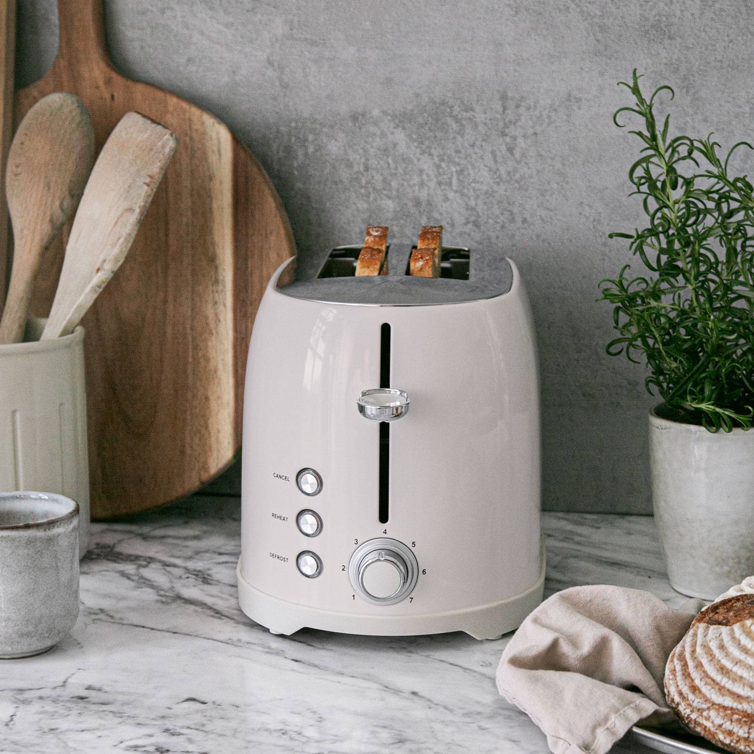 Een crèmekleurige broodrooster met toast staat op een marmeren aanrecht met keukengerei en een plant.