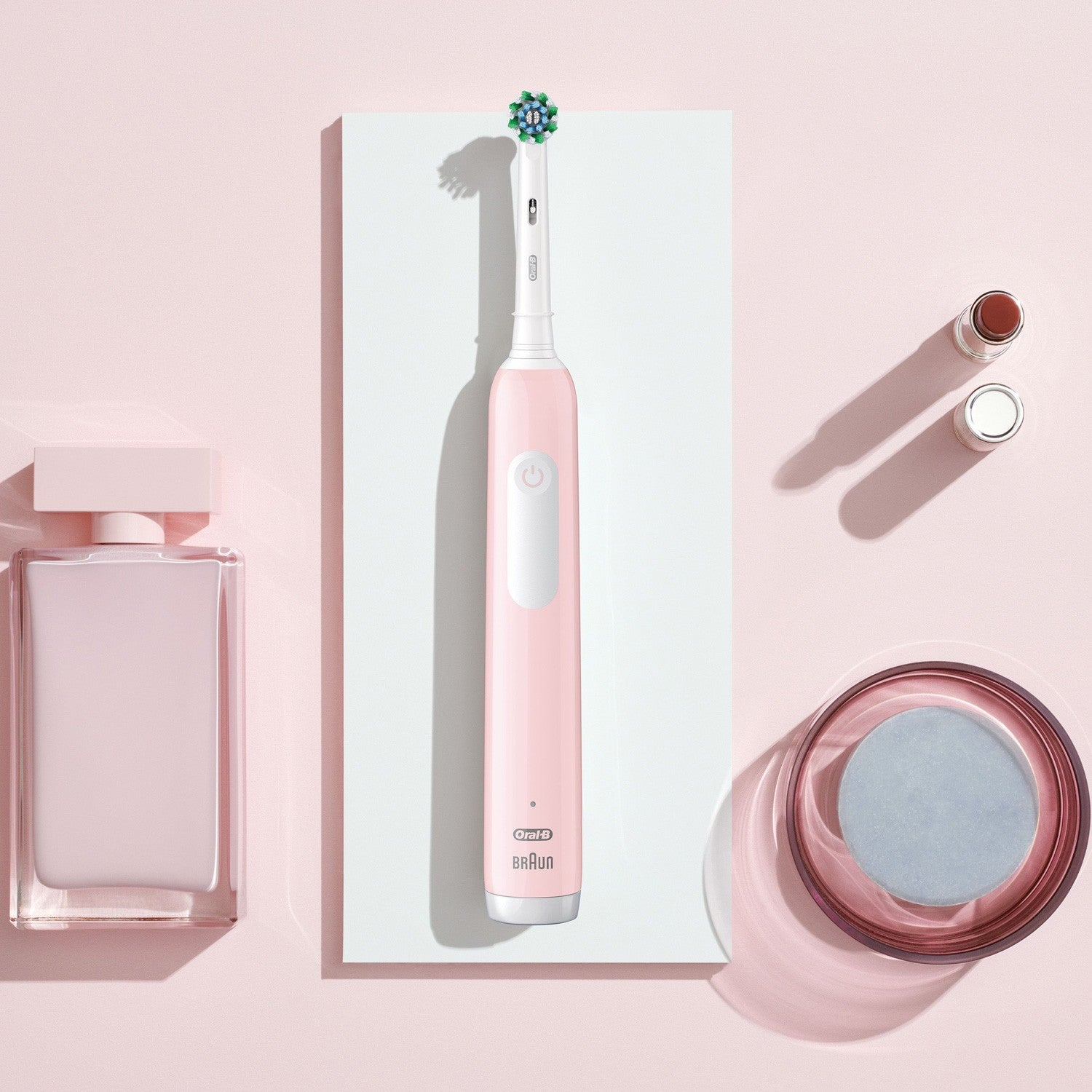 A pink electric toothbrush is on a white stand next to perfume and cosmetics.