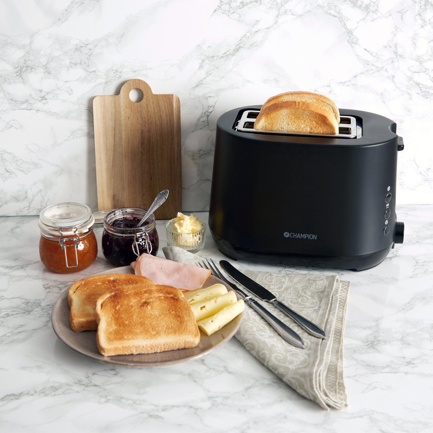 Zwarte broodrooster met toast, jam, boter, kaas en ham op marmer.