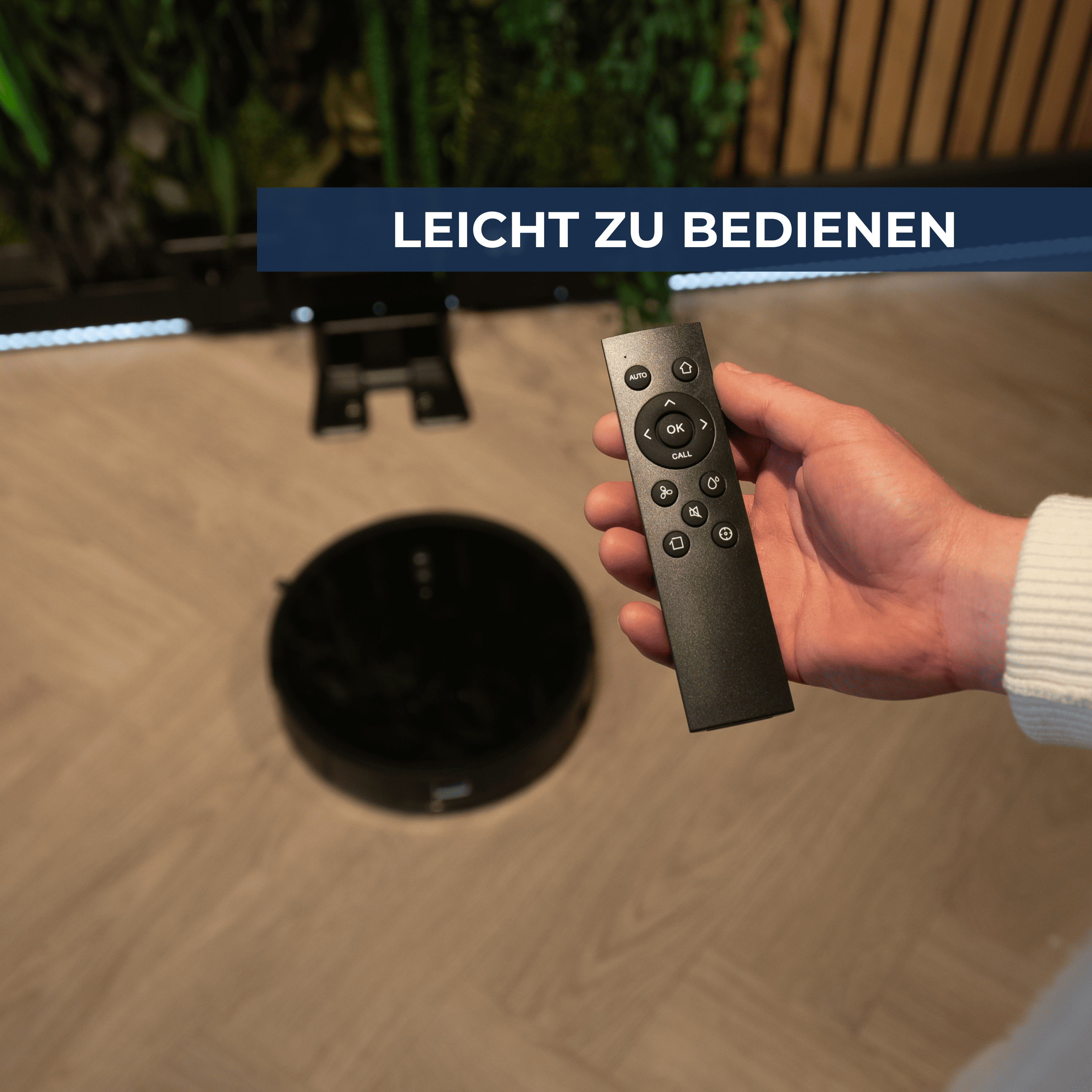 Eine Hand hält eine Fernbedienung neben einem runden Staubsaugerroboter.