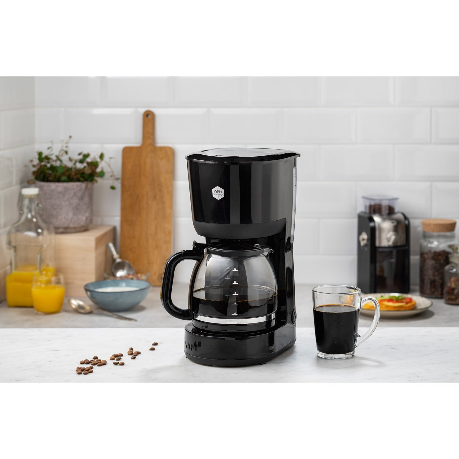 Cafetera negra con jarra de cristal y taza de café, junto a zumo de naranja y desayuno.