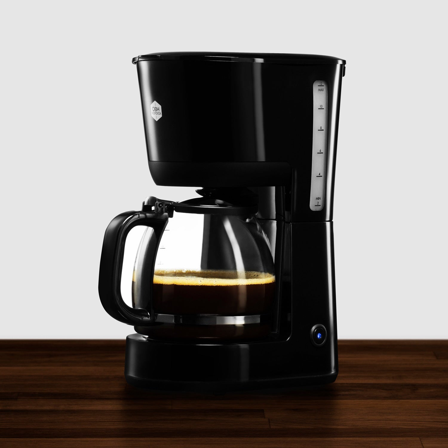 Cafetera negra con jarra de cristal, medio llena de café, sobre superficie de madera.