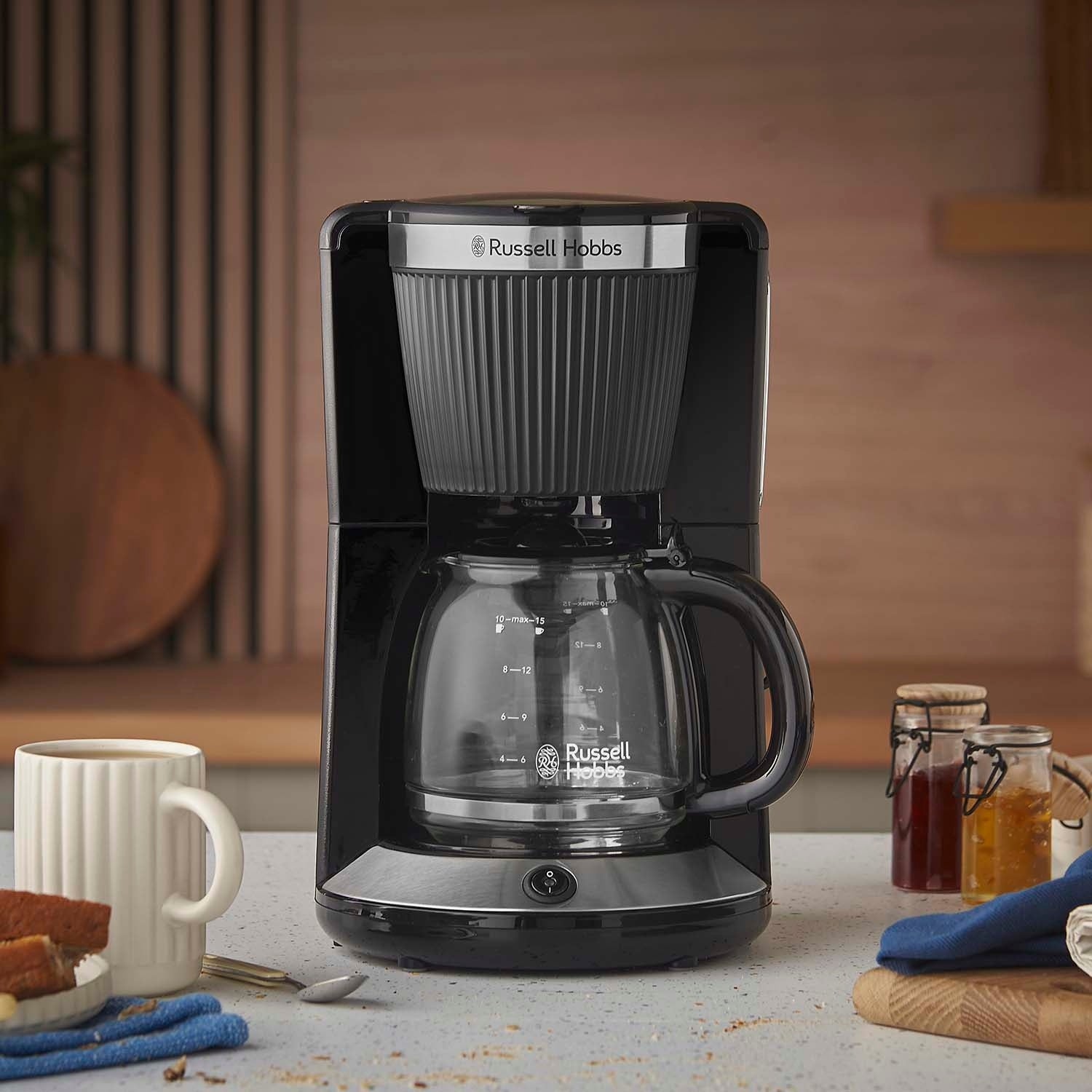 Cafetera Russell Hobbs negra con jarra de cristal, taza y pan en una encimera.