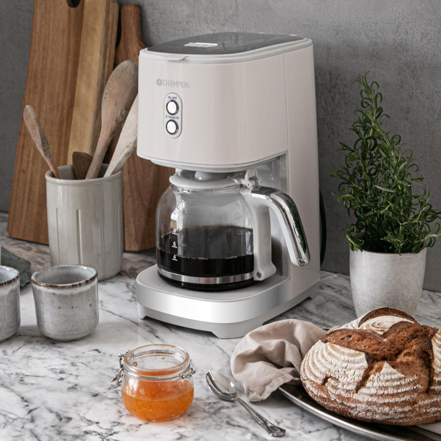 Una cafetera blanca con jarra de cristal llena de café, junto a pan, mermelada y tazas.