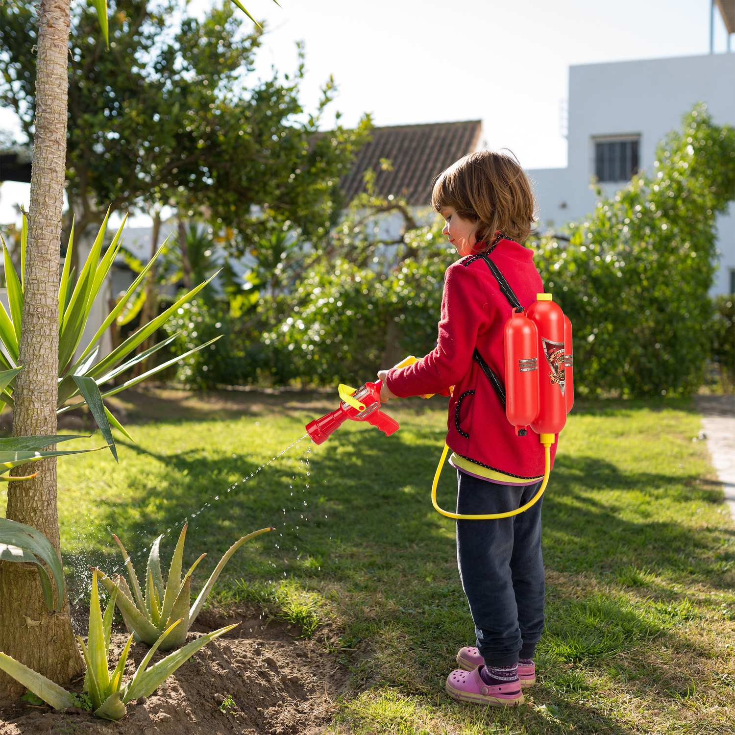 Un niño con mochila de agua roja rocía plantas de aloe en un jardín.