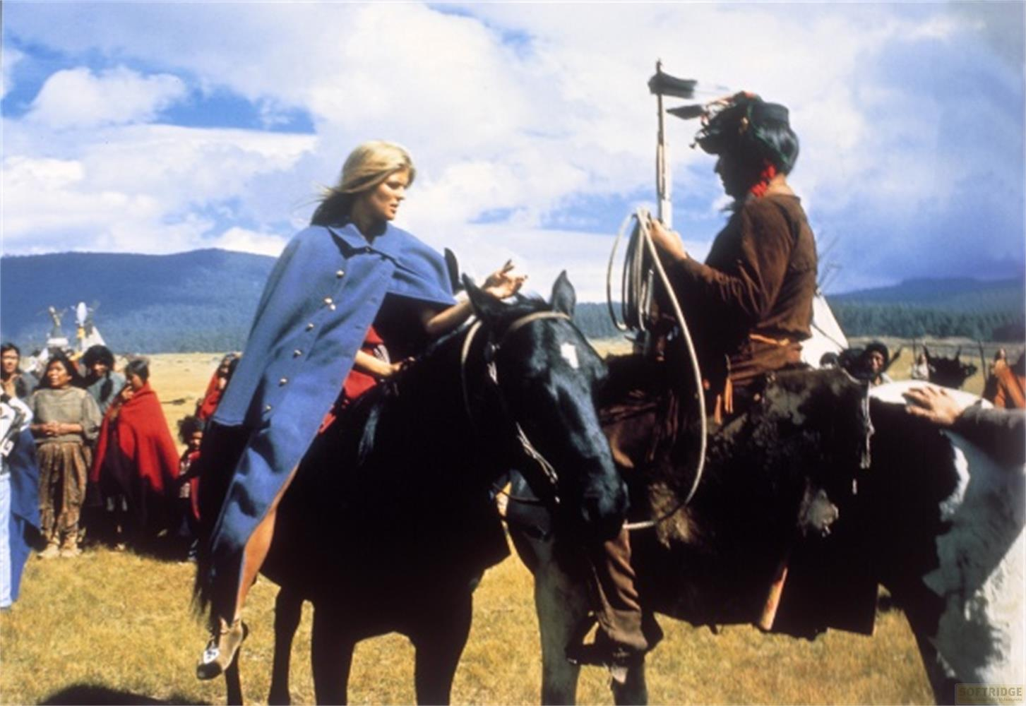 Una donna con mantello blu cavalca un cavallo nero, un uomo con un lazo cavalca accanto a lei.