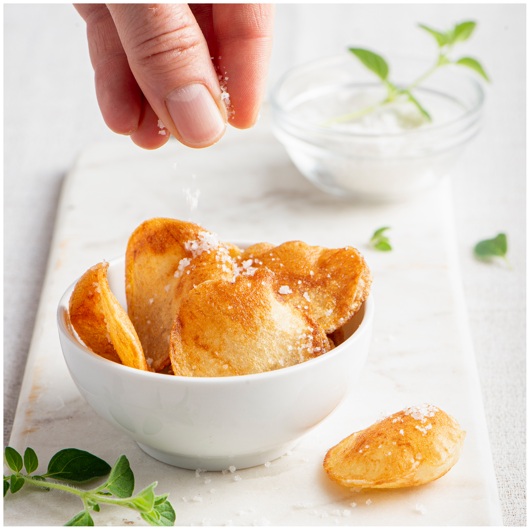 Een hand strooit zout over knapperige aardappelchips in een witte kom.
