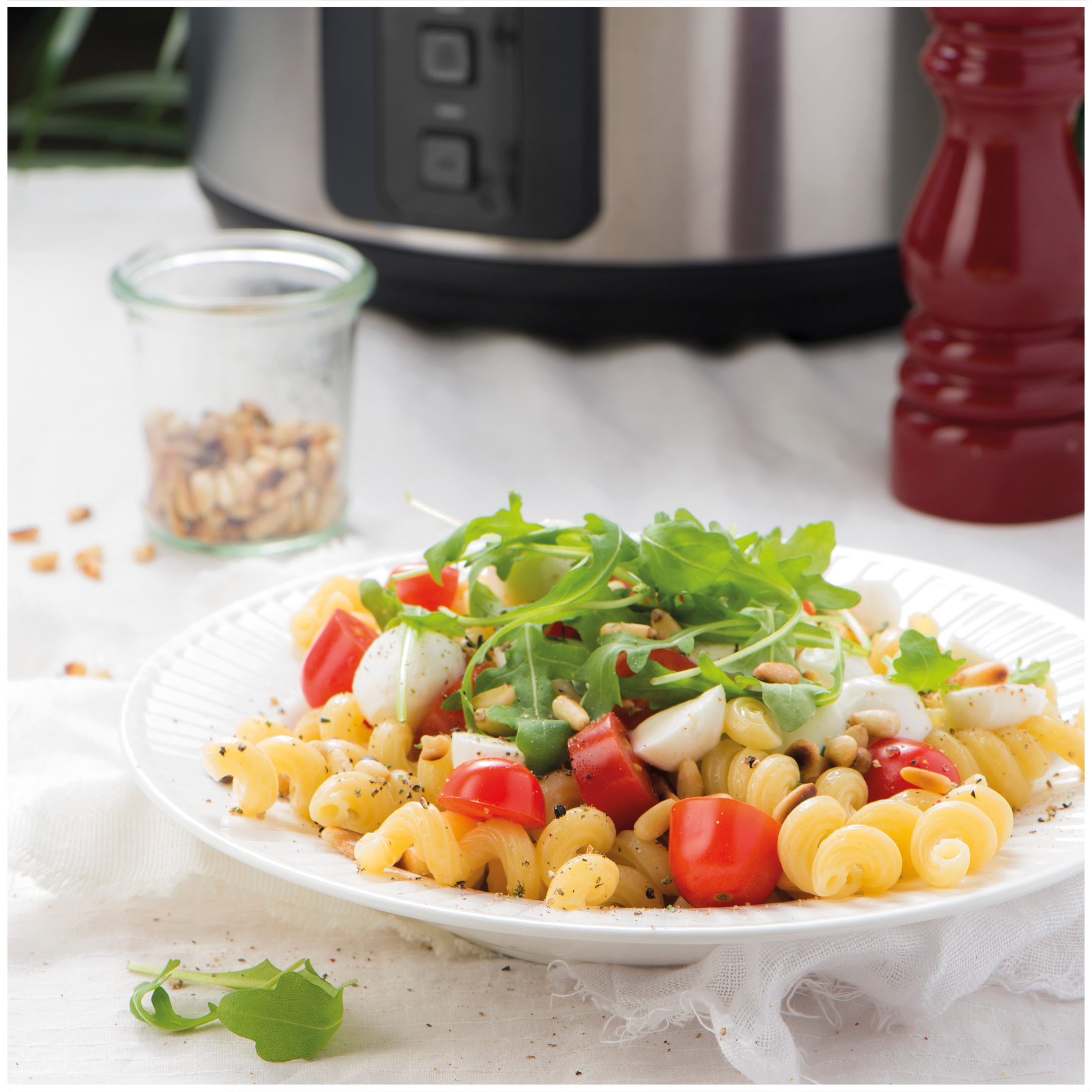 Pasta salad with cherry tomatoes, mozzarella balls, arugula, and pine nuts on a plate.