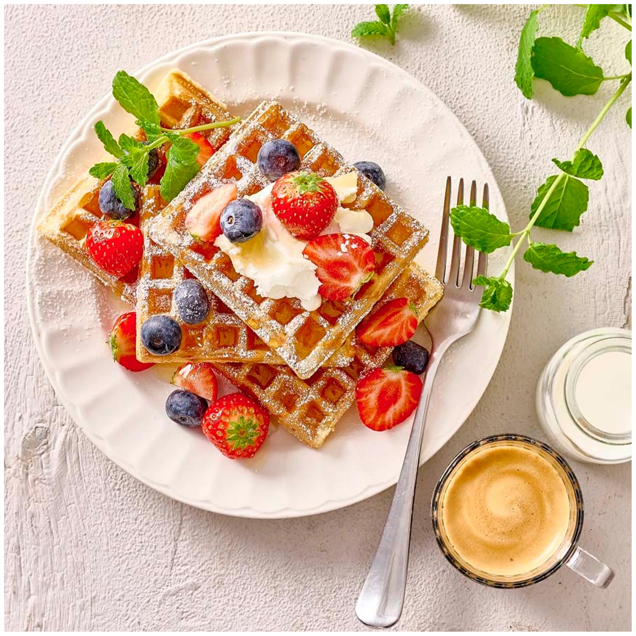 Waffle con frutti di bosco, panna e menta, accanto a caffè e latte.