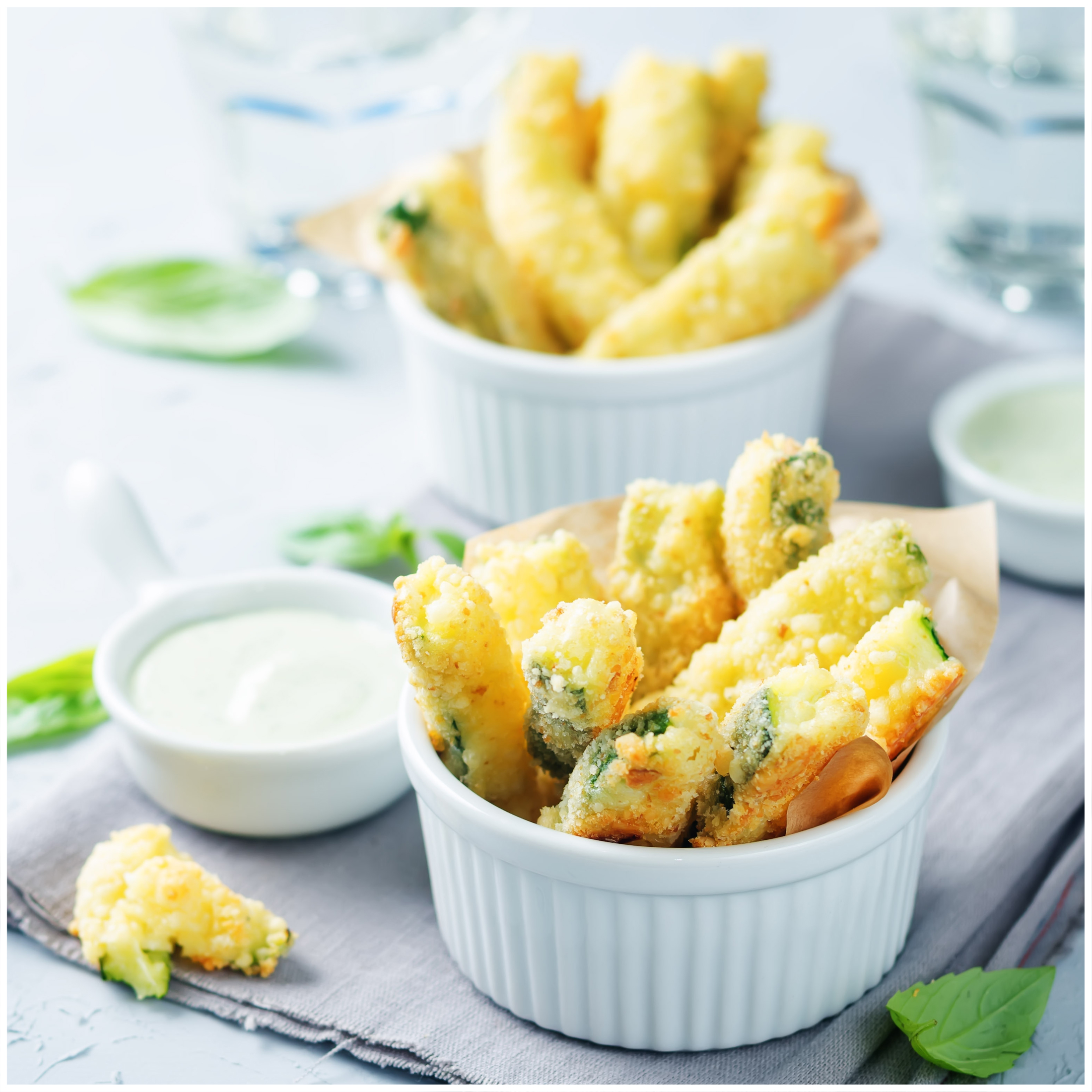 Gefrituurde courgette-sticks in witte schaaltjes met dipsaus en basilicum.