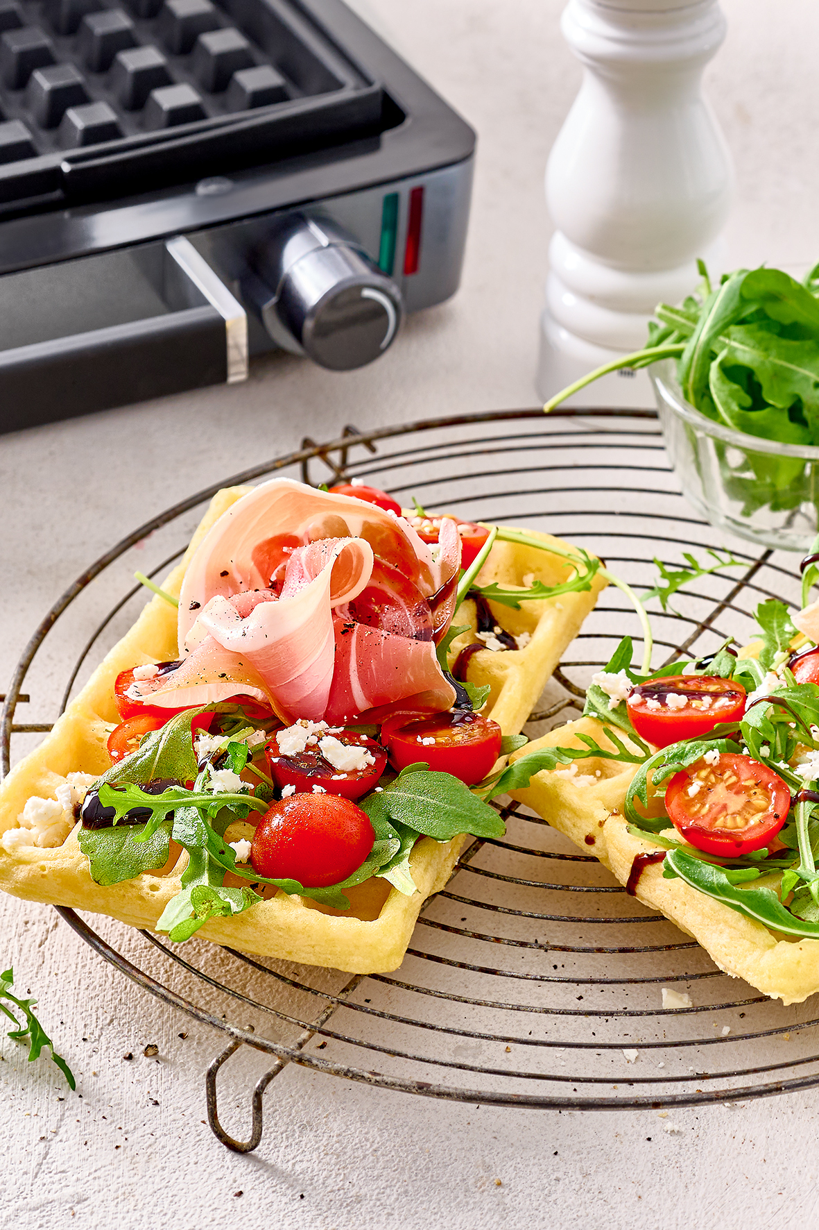 Waffle conditi con prosciutto, pomodorini, feta e rucola su una gratella.