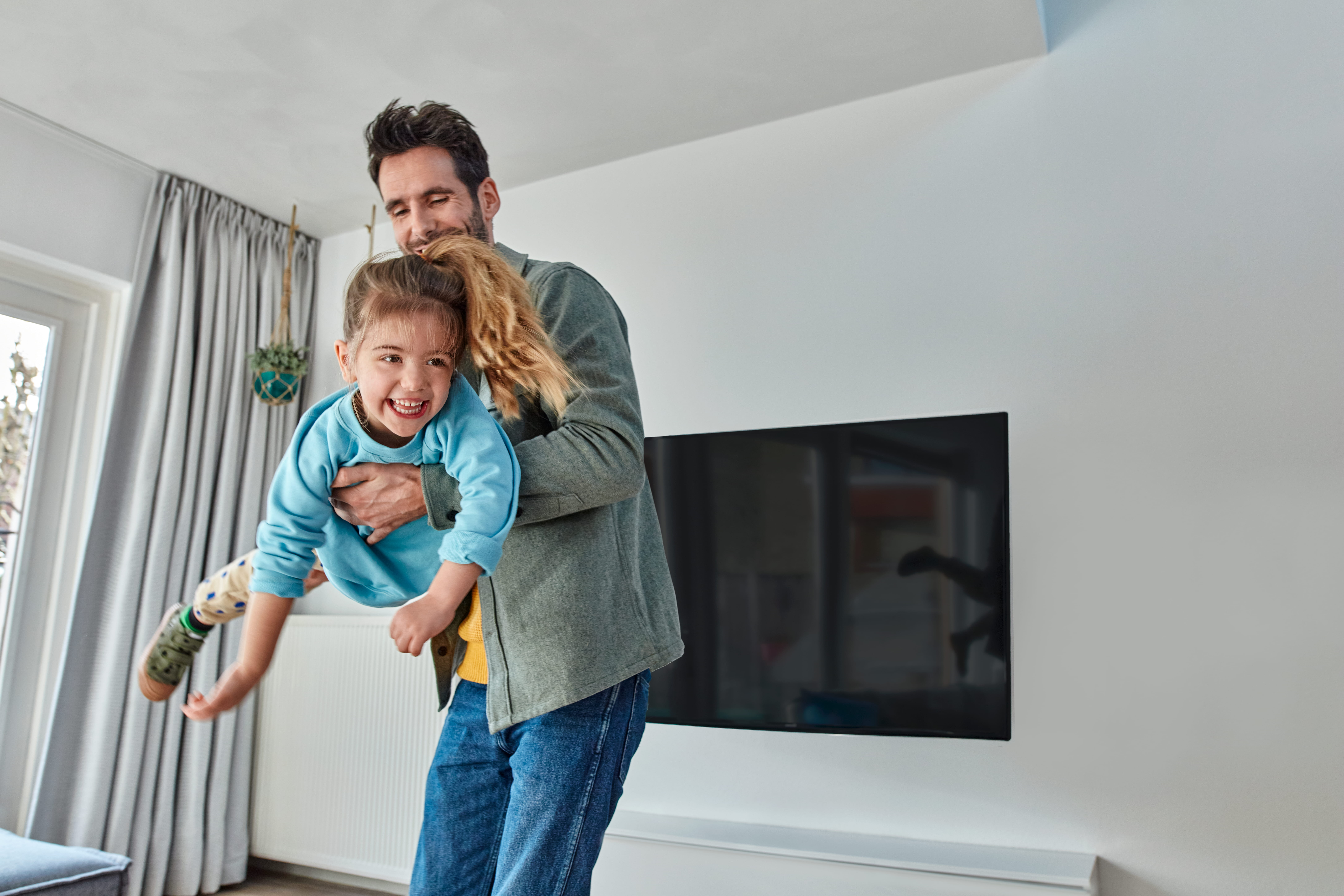 Ojciec trzyma córkę na rękach, śmieją się w salonie.