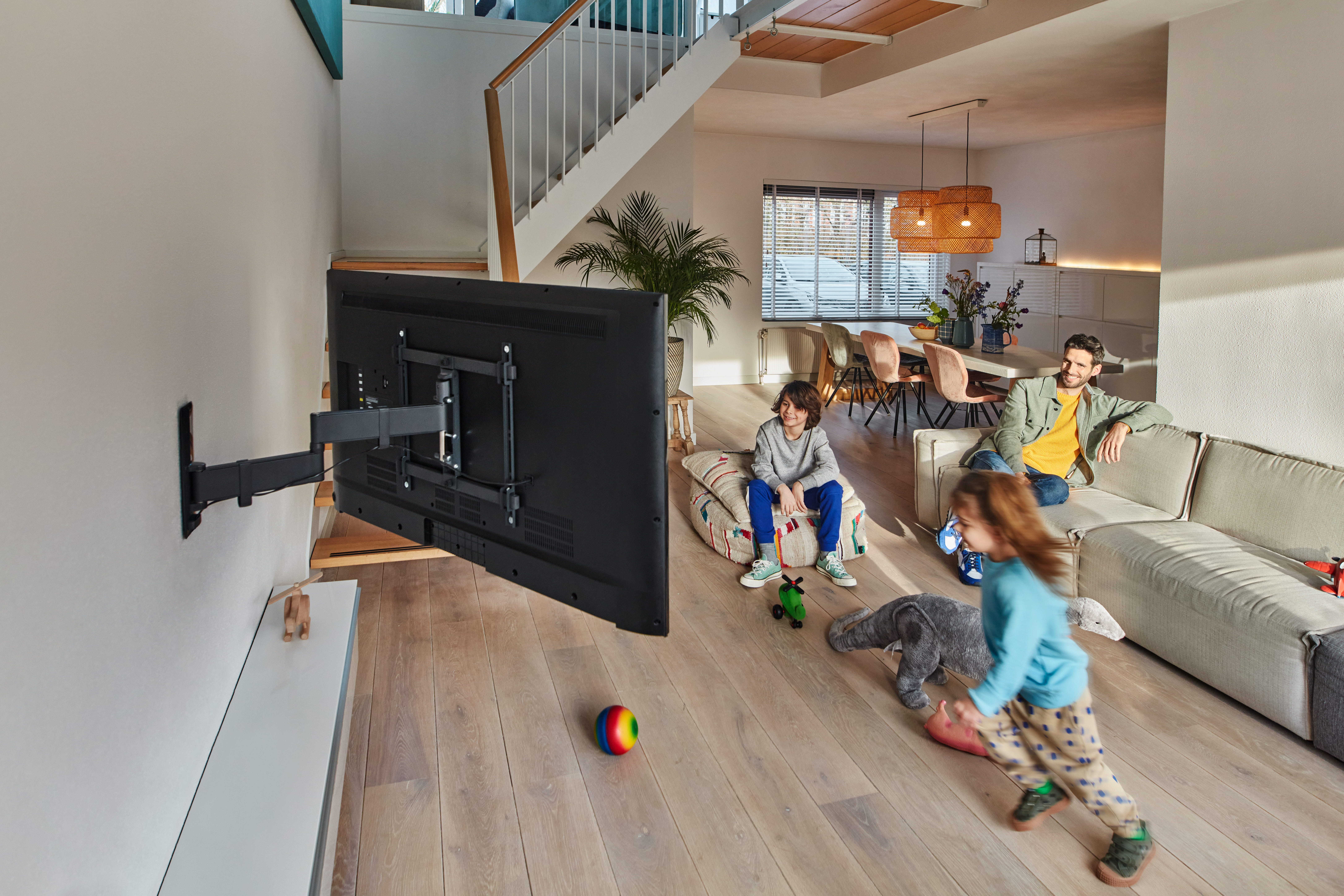 Rodzina w salonie: mężczyzna na kanapie, dzieci bawią się na podłodze, TV na ścianie.