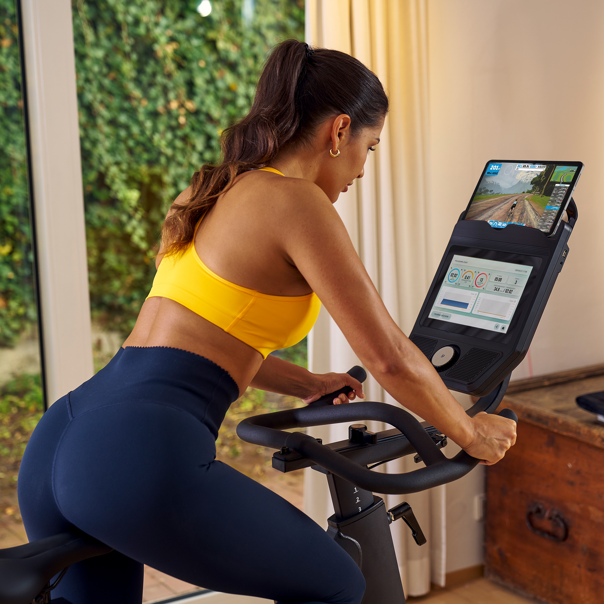 Frau auf Heimtrainer vor Bildschirm mit Radrennspiel, gelbes Top, blaue Hose.