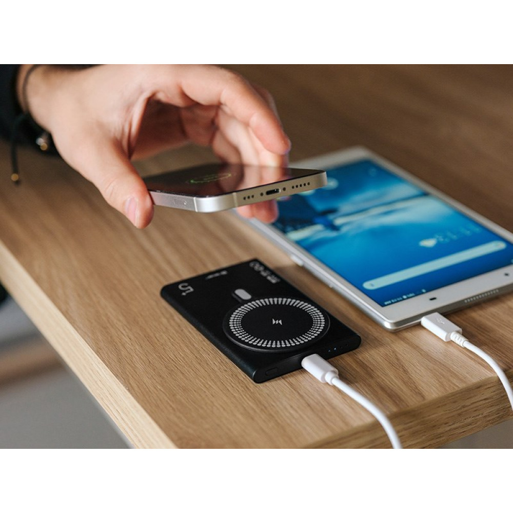 Dłoń trzyma telefon nad czarną ładowarką bezprzewodową obok tabletu.