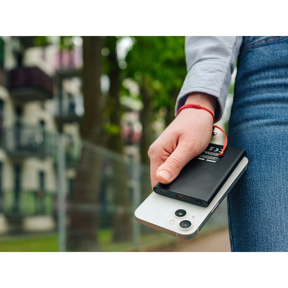 Ręka trzyma czarną powerbank na białym smartfonie. W tle budynki.