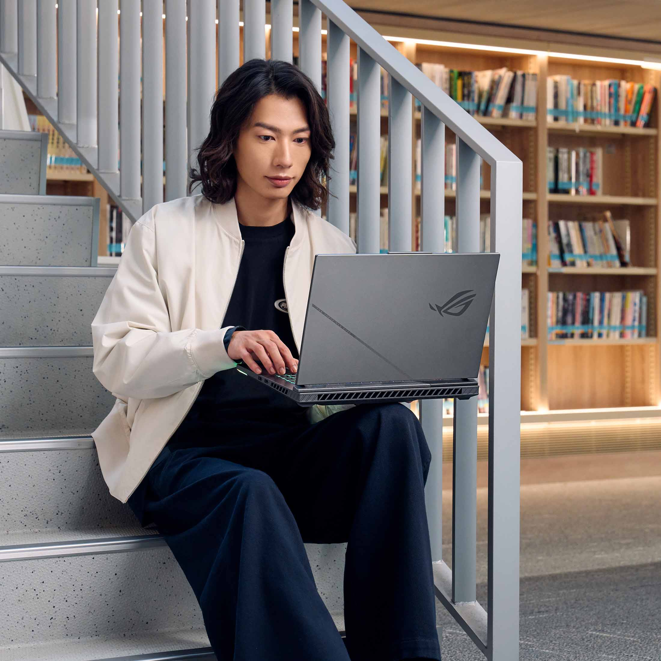 Ein Mann sitzt auf einer Treppe und benutzt einen Laptop. Im Hintergrund sind Regale mit Büchern.